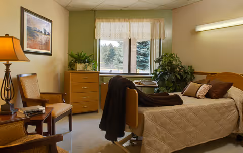 A furnished bedroom in a senior living facility with a bed, dresser, and plants.