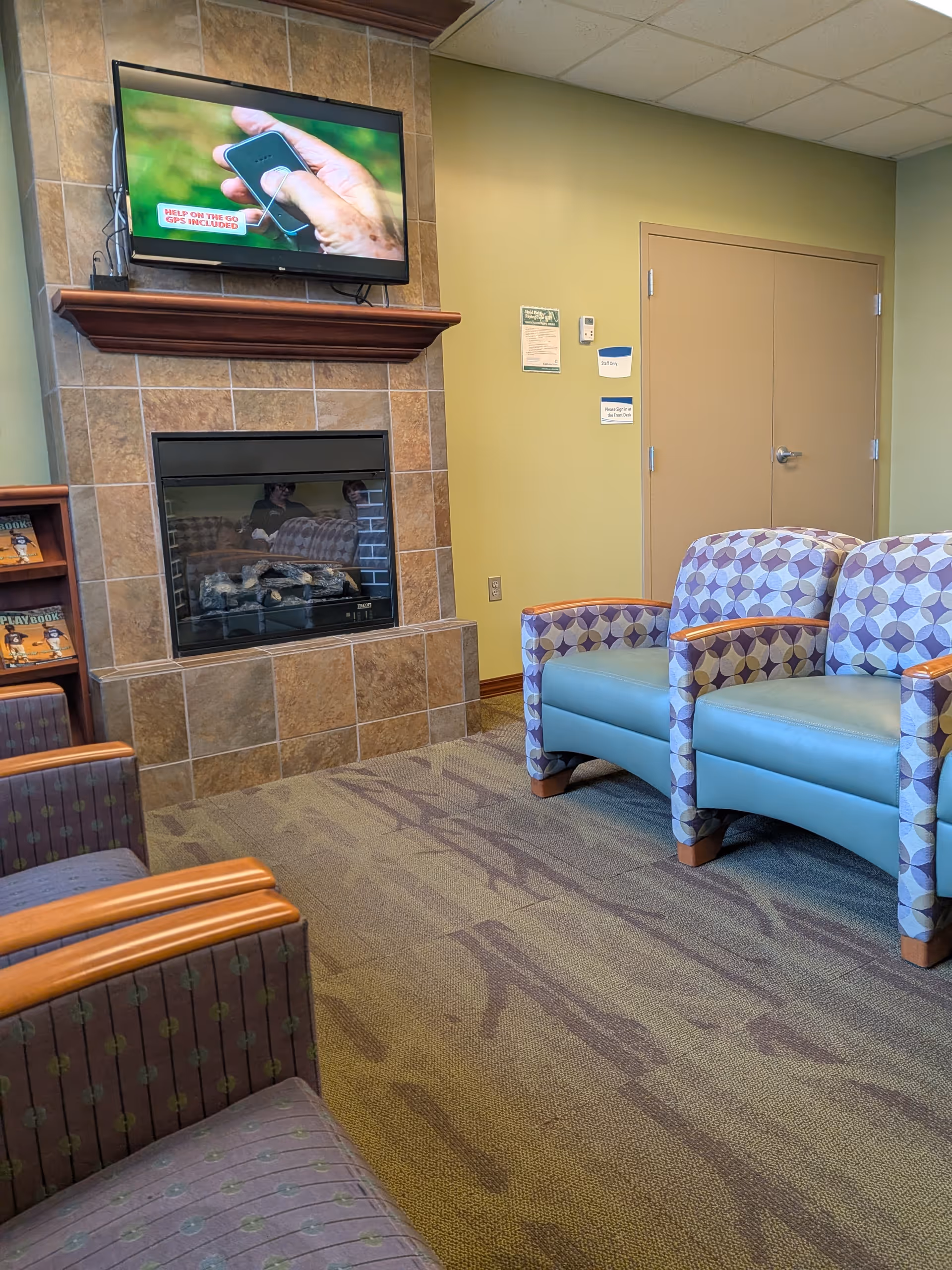 Cozy resident lounge featuring a tiled fireplace, wall-mounted TV, and patterned blue chairs.
