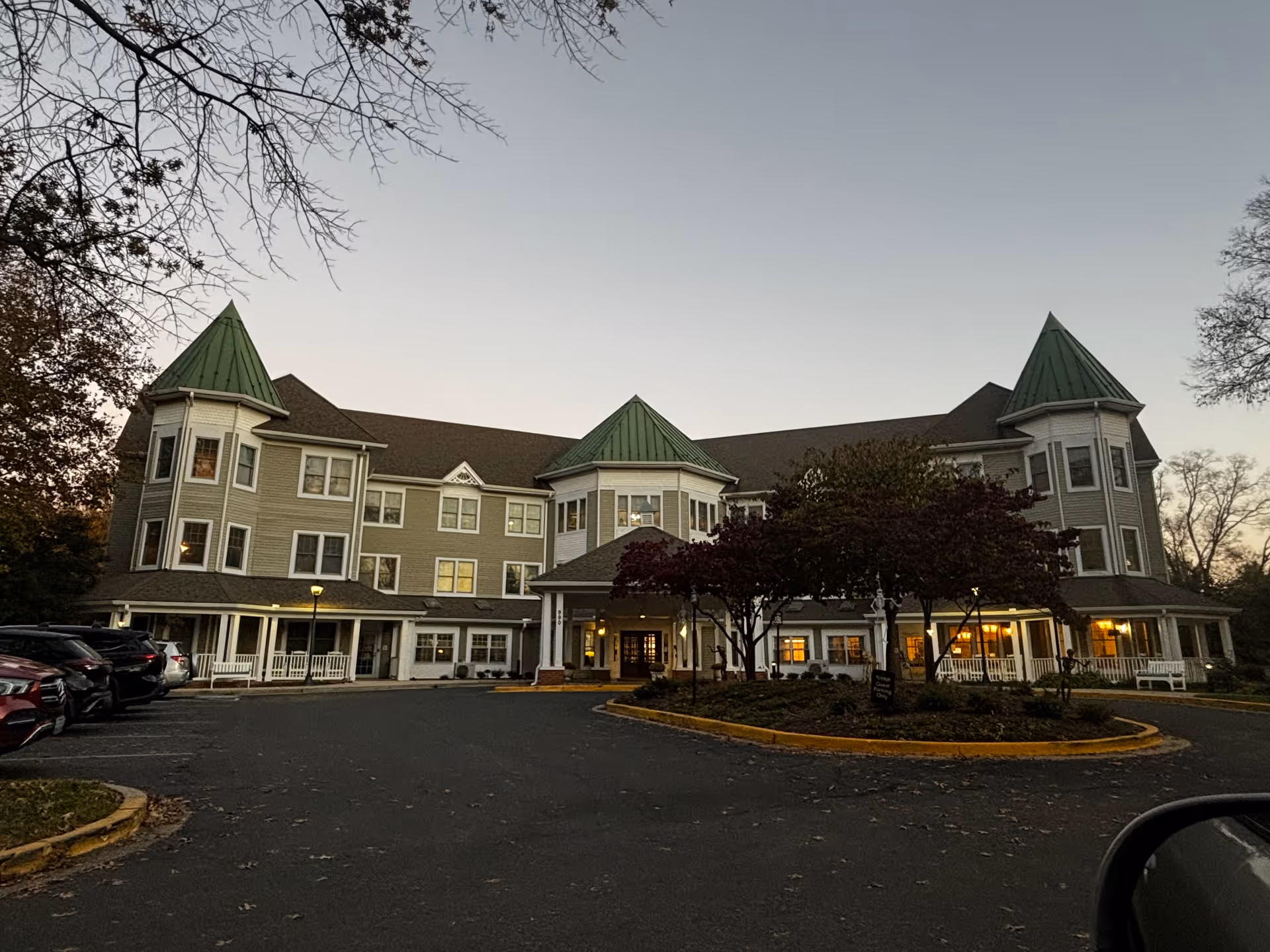 Exterior view of a large senior living facility building with green pointed roofs and multiple windows, surrounded by trees and a parking lot with several cars parked. The sky is clear and it appears to be either early morning or late evening.