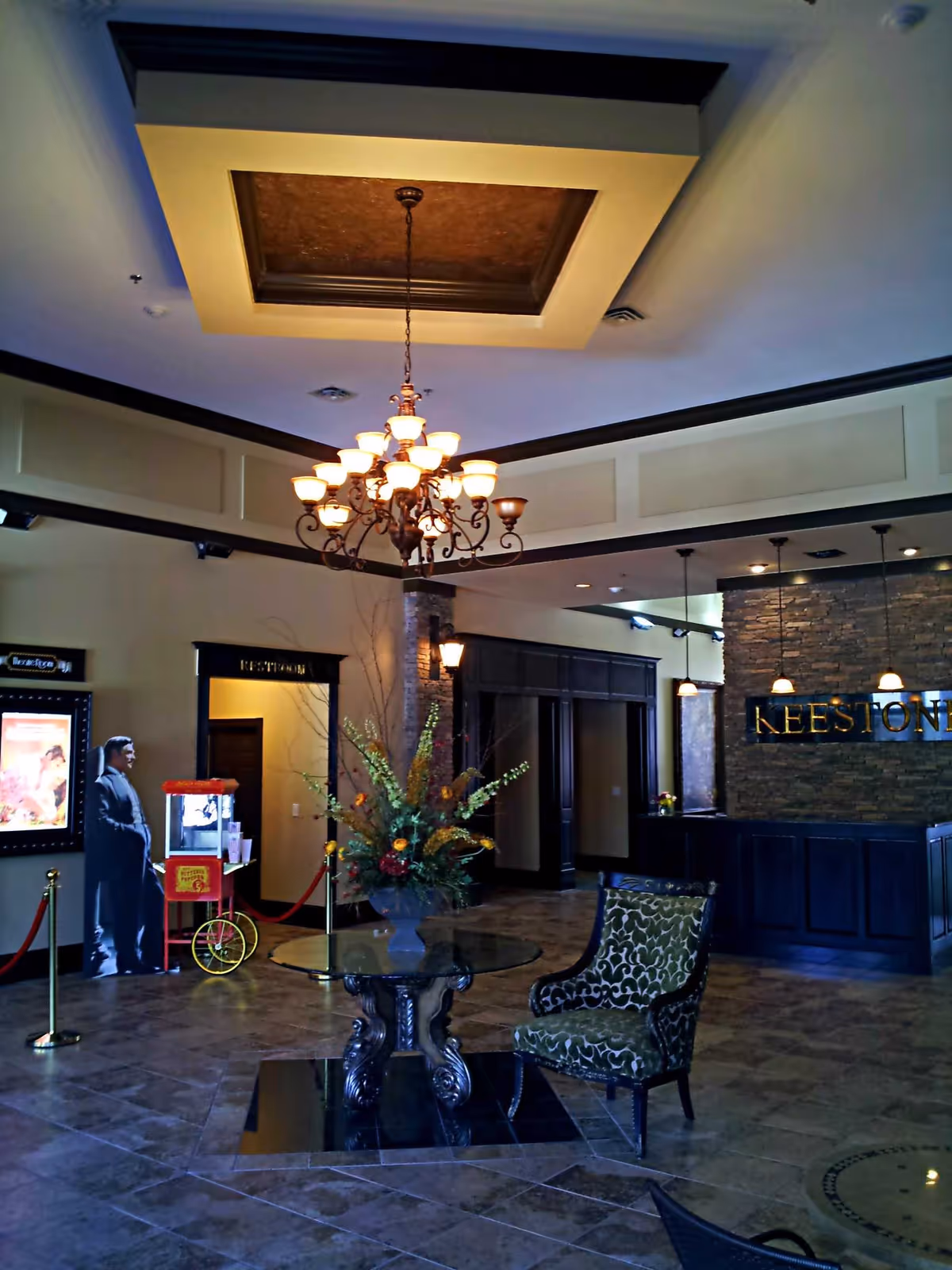 Interior view of a senior living facility lobby with a decorative chandelier hanging from the ceiling, a round glass table with a large floral arrangement, a patterned upholstered chair, and a reception desk with the word 'KEESTONE' on the stone wall behind it. There is also a popcorn machine and a life-size cutout of a person near a doorway labeled 'Restrooms'.