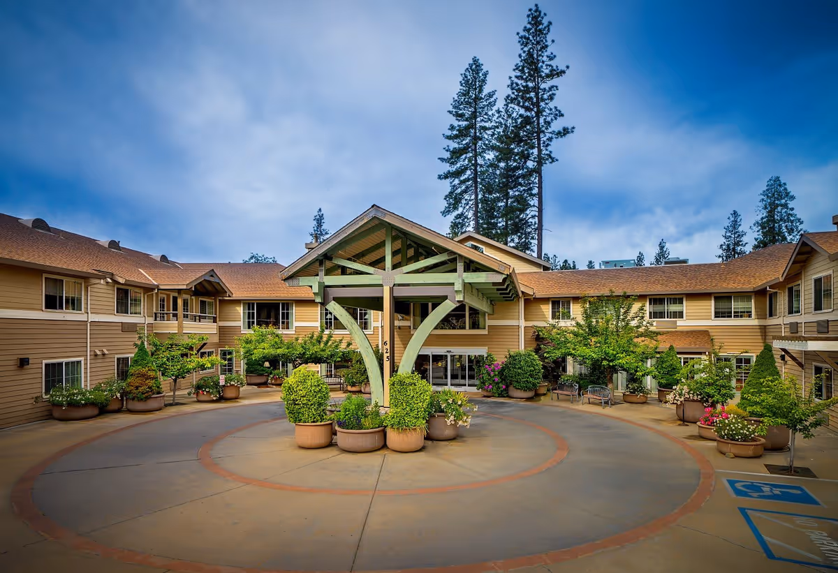 Front exterior view of Eskaton Village Grass Valley showing a circular driveway with a covered entrance supported by green wooden beams, surrounded by potted plants and trees, with a two-story beige building and tall pine trees in the background under a cloudy sky.
