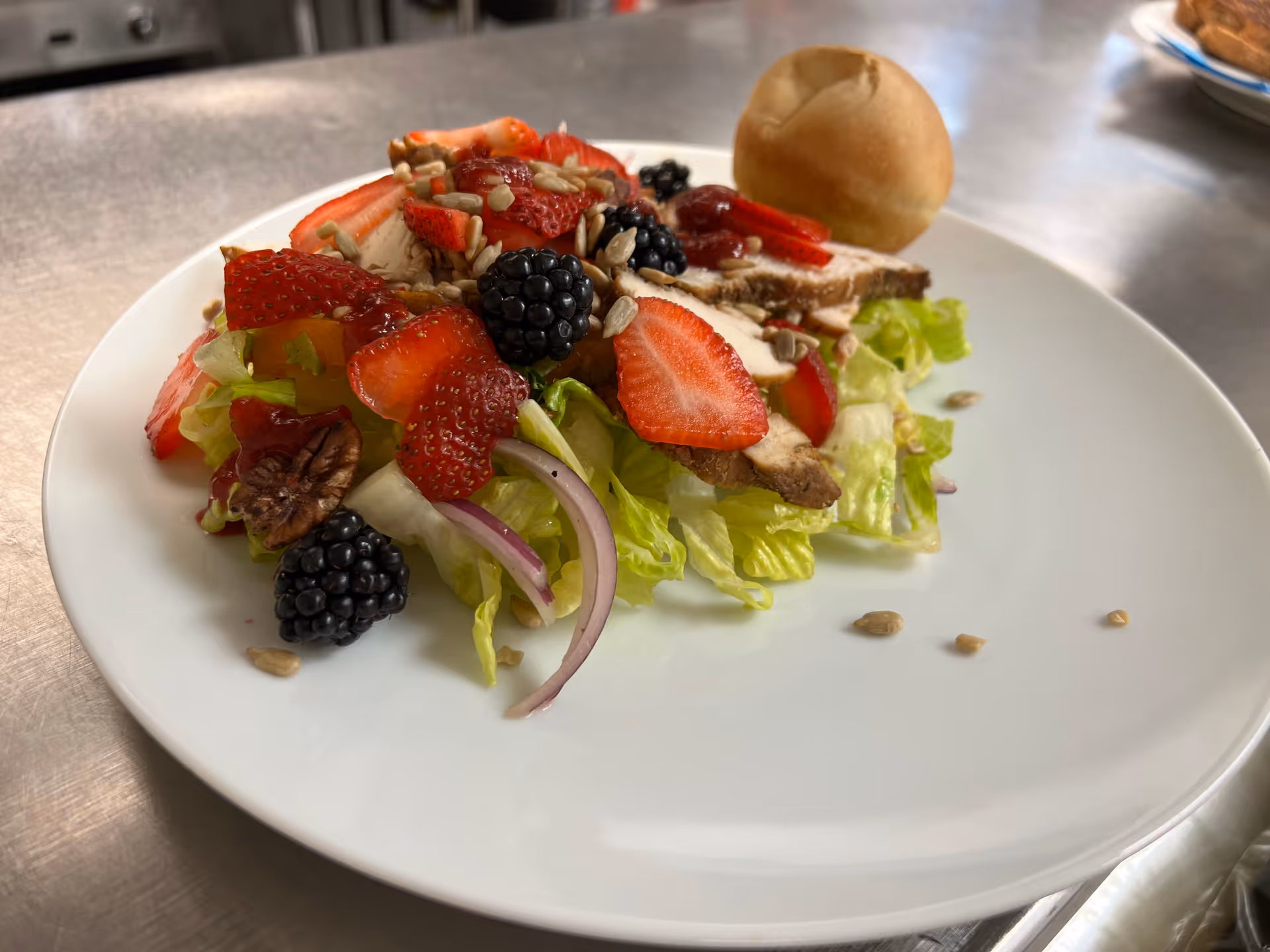 A plate with a fresh salad consisting of lettuce, sliced strawberries, blackberries, pecans, red onion slices, sunflower seeds, and grilled chicken slices topped with a dressing. A small bread roll is placed on the side of the plate.
