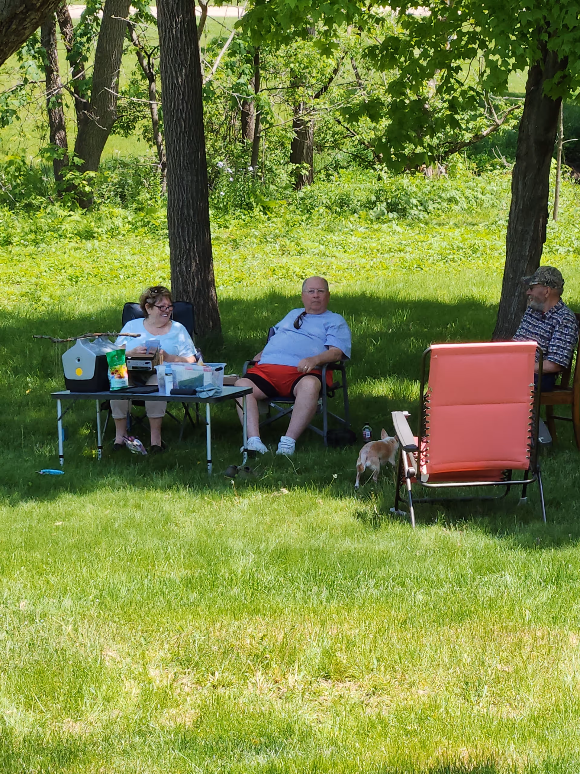 Three seniors sit on lawn chairs under trees beside a table with supplies outdoors.
