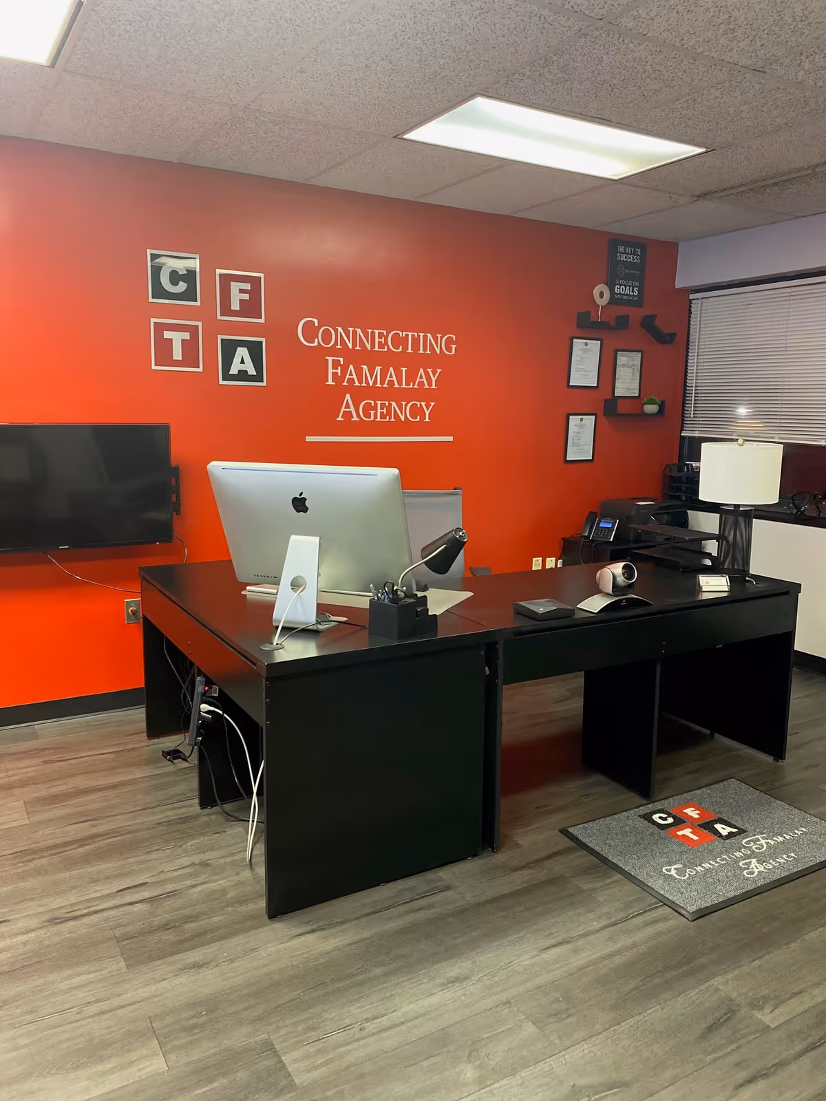 Office reception with red wall, desk, computer monitor, and Connecting Famalay Agency signage.
