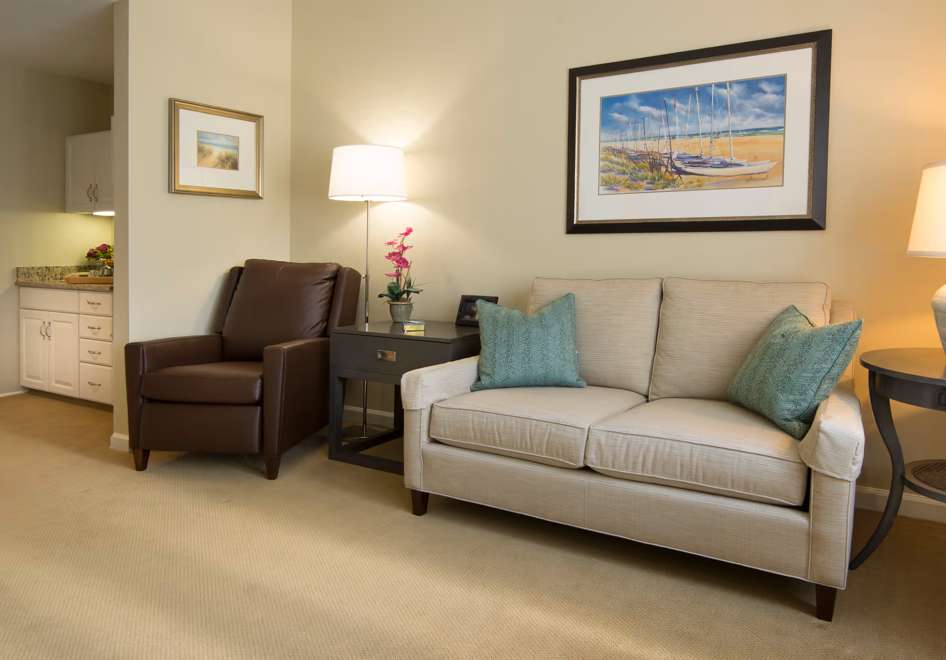 A cozy living room with a beige sofa topped with teal pillows, a brown leather armchair, side tables and lamps, and framed artwork on the wall.