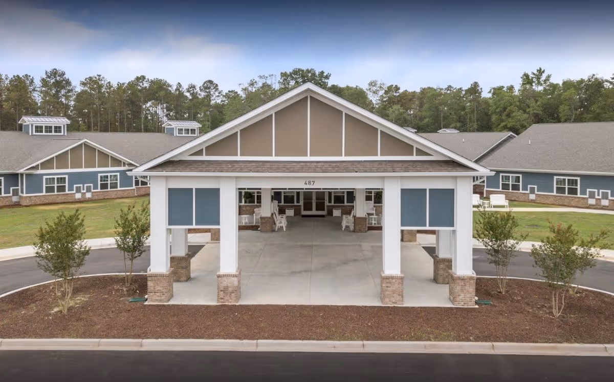Covered front entrance (porte-cochere) of a single-story senior living building with white columns, blue siding, and surrounding landscaping.