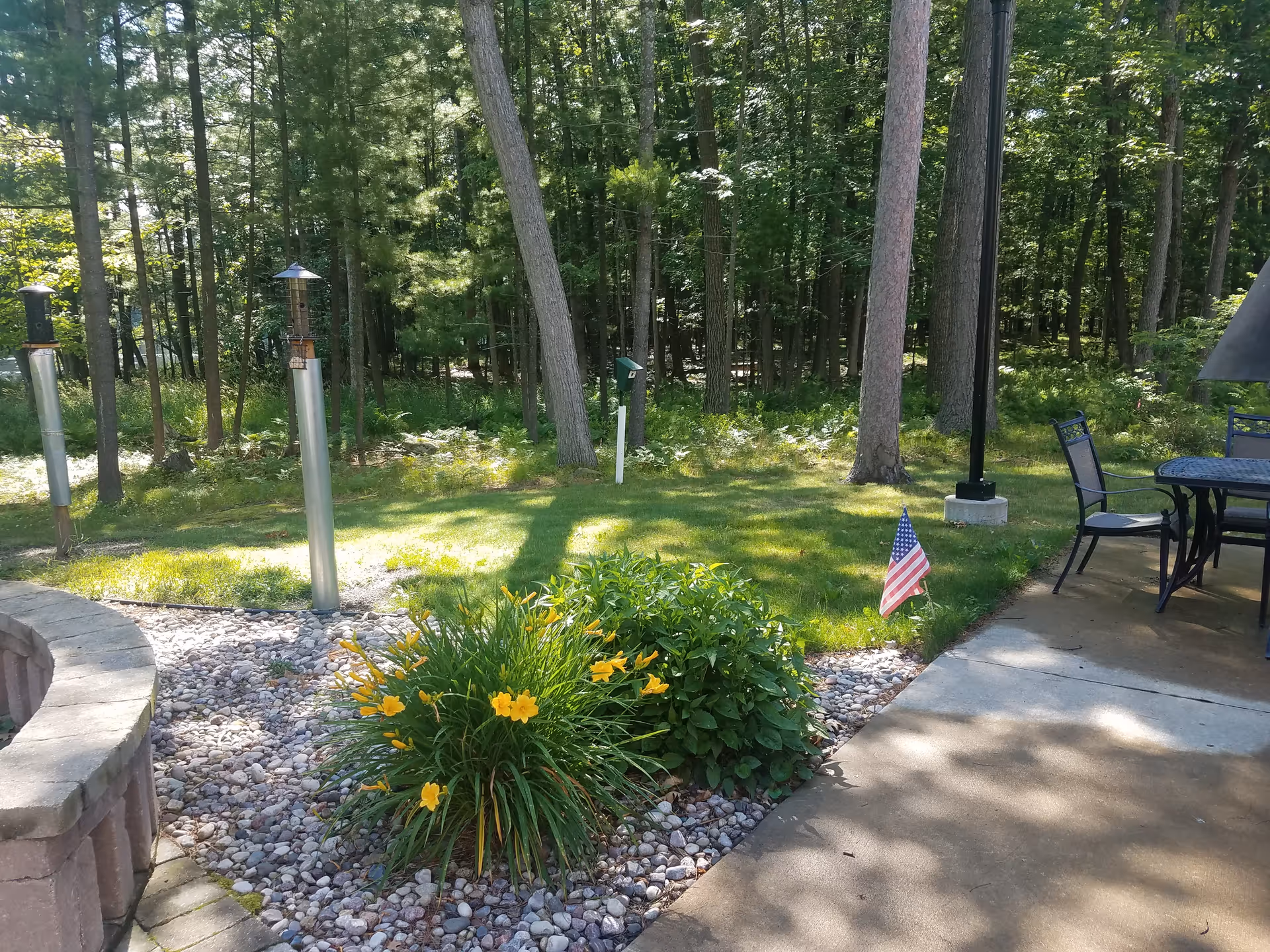 Outdoor patio area with metal table and chairs, flag, plant bed, and trees.