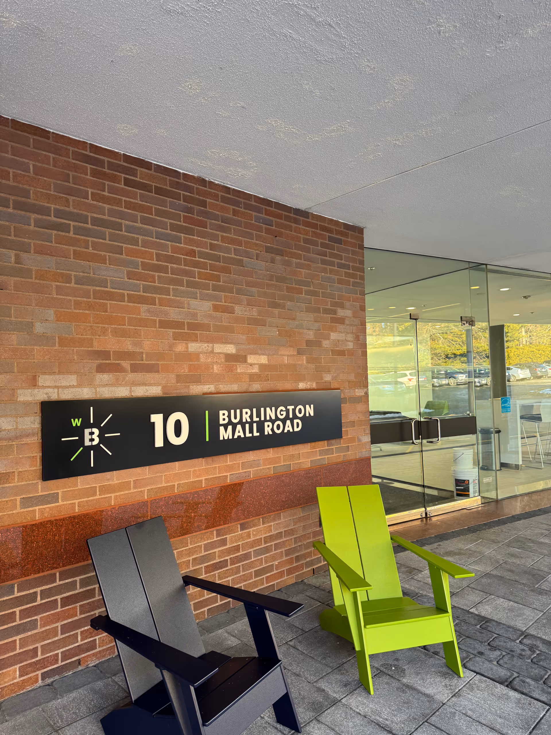 Two chairs, black and bright green, sit outside a brick building with a glass entrance.
