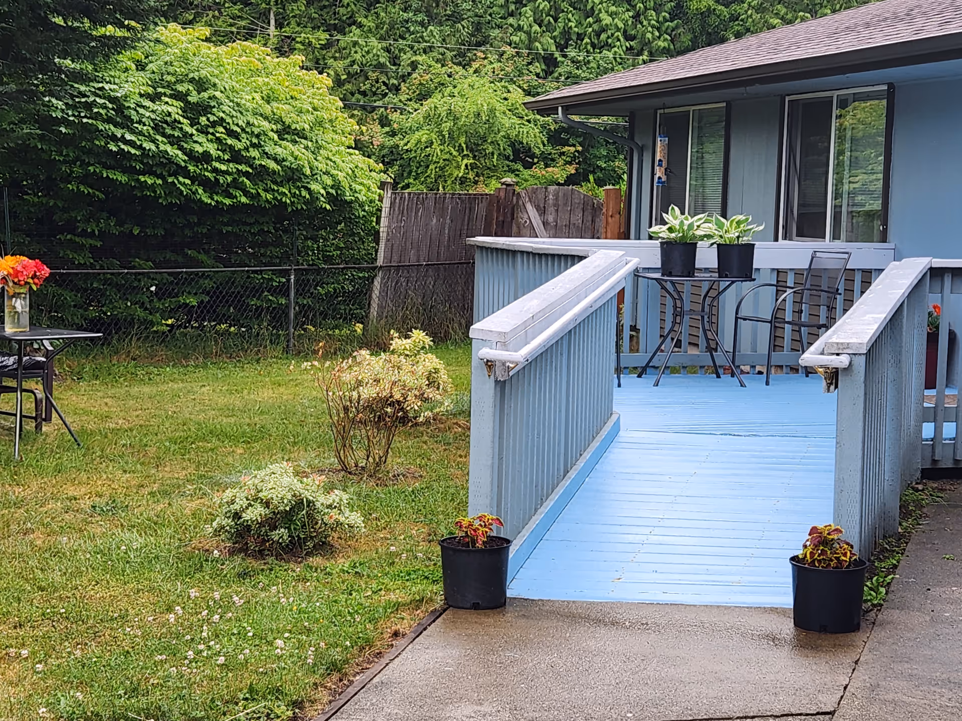 Blue wooden deck with ramp, railings, potted plants, and patio furniture beside a blue house.