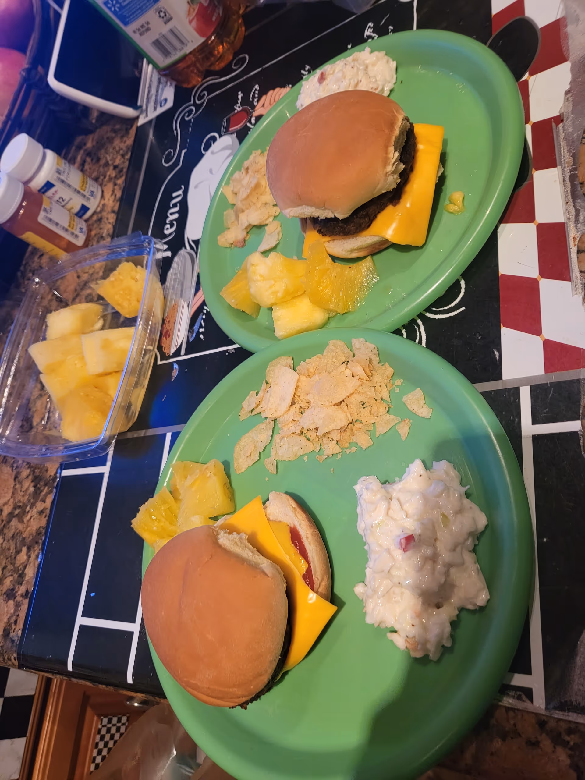 Two green plates with burgers, cheese, pineapple, and side dishes on a dining table.