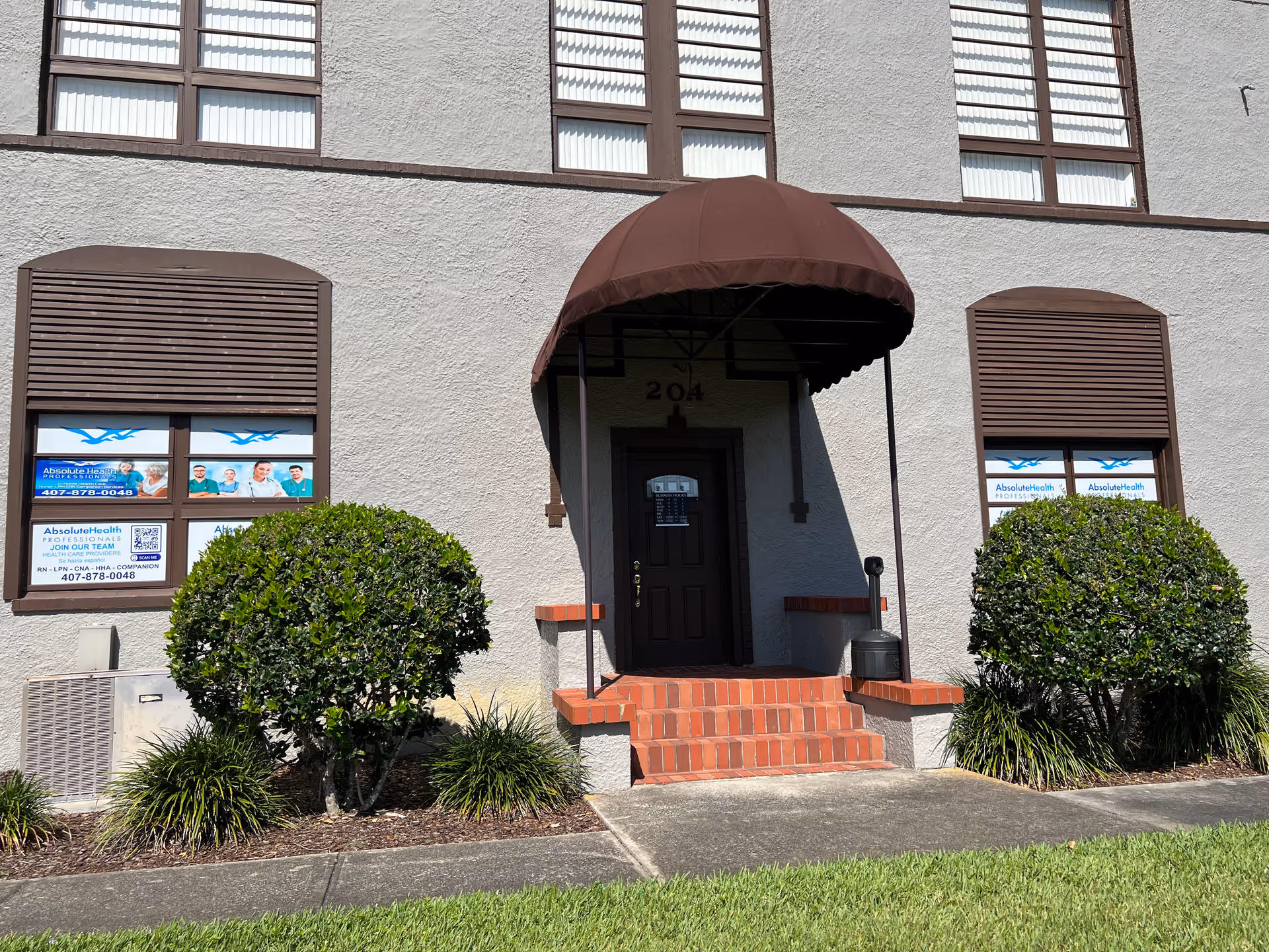 Front entrance of a beige building with a brown canopy, red brick steps, and shrubs.