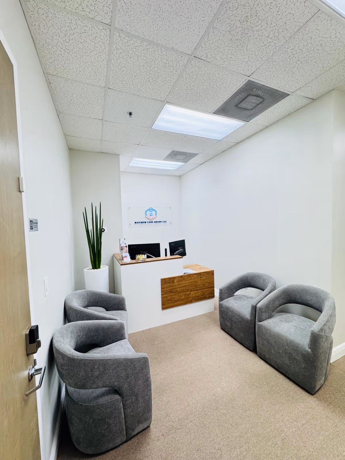 Small reception area with gray curved chairs, a white desk, and a plant near the doorway.