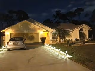 Night view of a single-story residential building with a driveway and a parked car. The house is illuminated by outdoor lights along the driveway and near the entrance. There are some bushes and trees around the house.