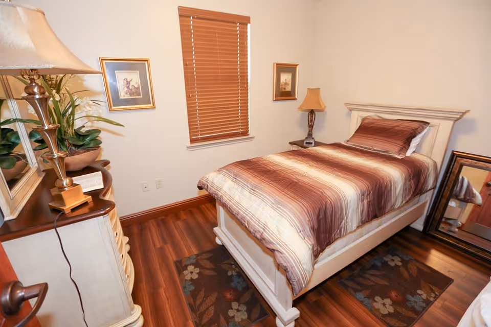 A small bedroom with a single bed featuring a striped brown and beige comforter. The room has wooden flooring with two floral rugs, a window with wooden blinds, a dresser with a lamp and a plant, two framed pictures on the walls, and a large mirror resting against the wall.