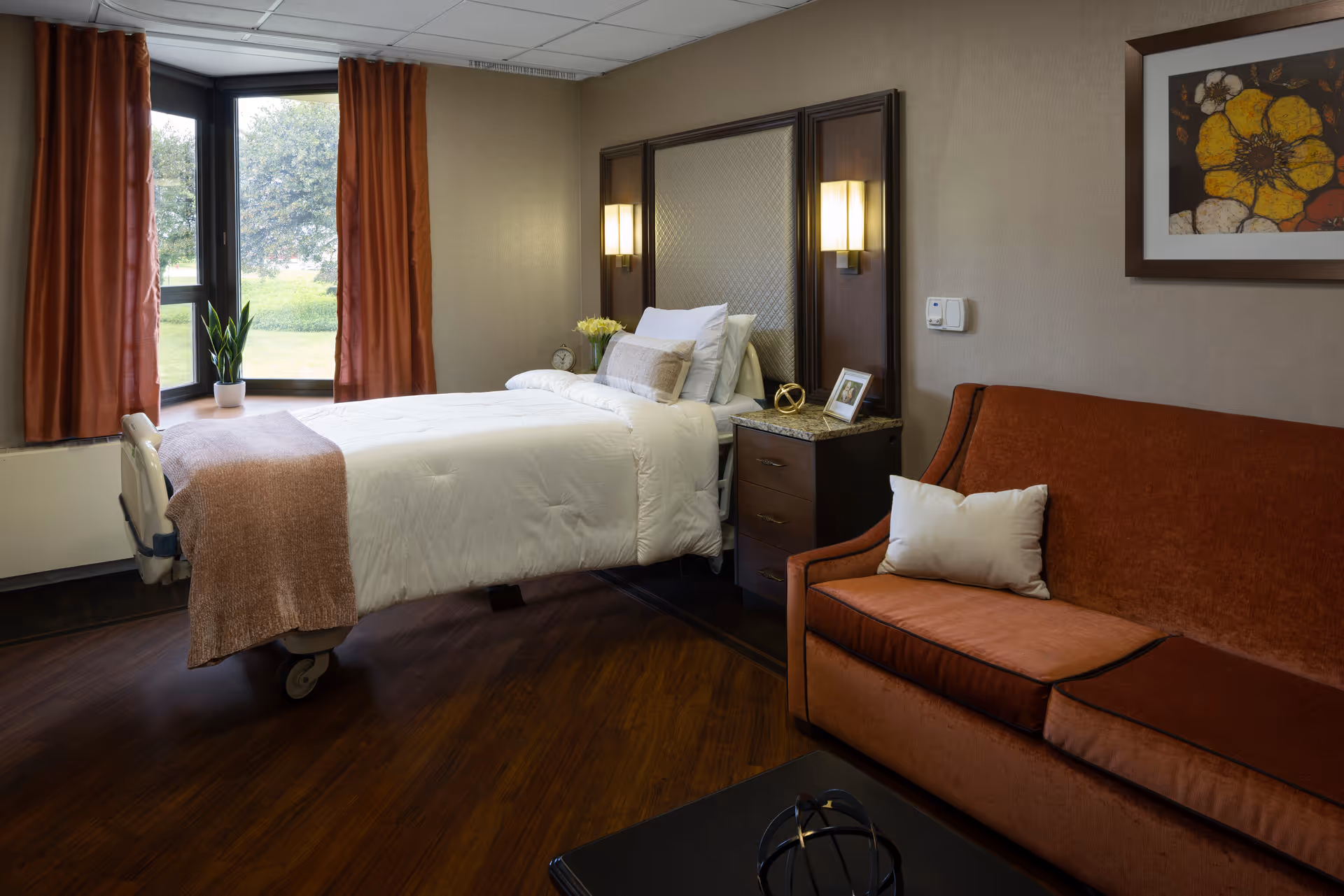 A cozy bedroom in a senior living facility featuring a single bed with white bedding and a light brown throw blanket. The bed is positioned near a window with rust-colored curtains and a small potted plant on the windowsill. Next to the bed is a dark wooden nightstand with a small clock, a framed photo, and decorative items. On the right side of the room is a rust-colored sofa with a white pillow, and above it hangs a framed floral artwork. The room has wooden flooring and soft lighting from wall-mounted lamps.