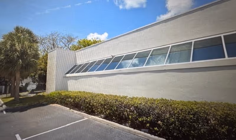 Exterior view of a modern building with slanted long windows, hedges, and a parking lot.