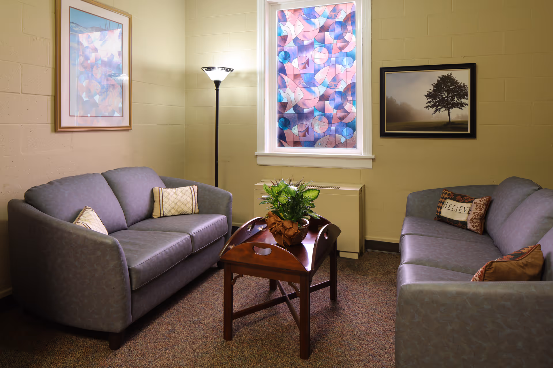 A small sitting area with two gray sofas facing each other, each with decorative pillows. Between them is a wooden coffee table with a potted plant. The room has beige walls with two framed pictures, one abstract and one of a tree, and a tall floor lamp in the corner. A window with colorful stained glass is centered on the back wall.