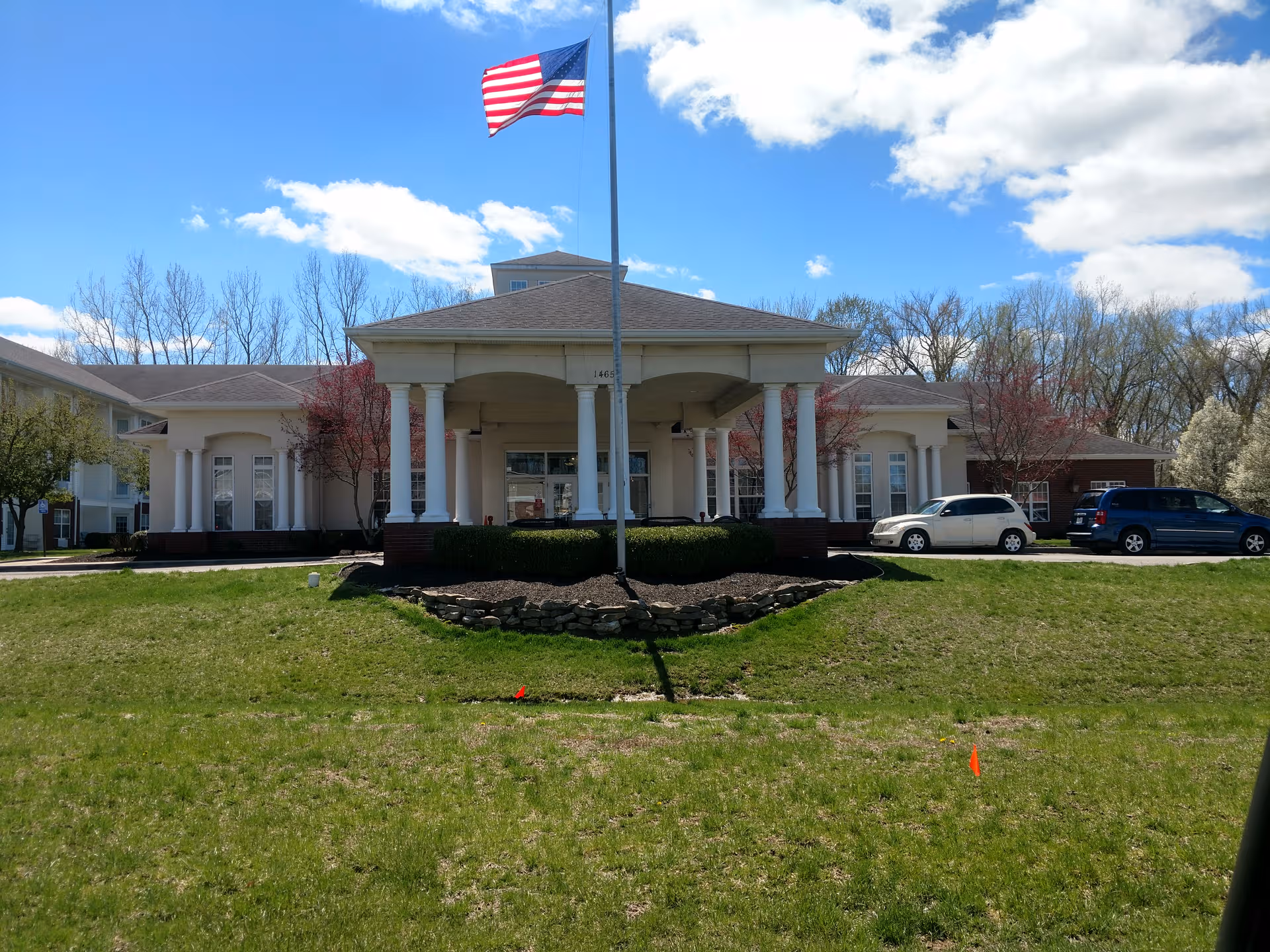 Front view of Wyndmoor Assisted Living entrance with columns, flagpole, and parked cars.