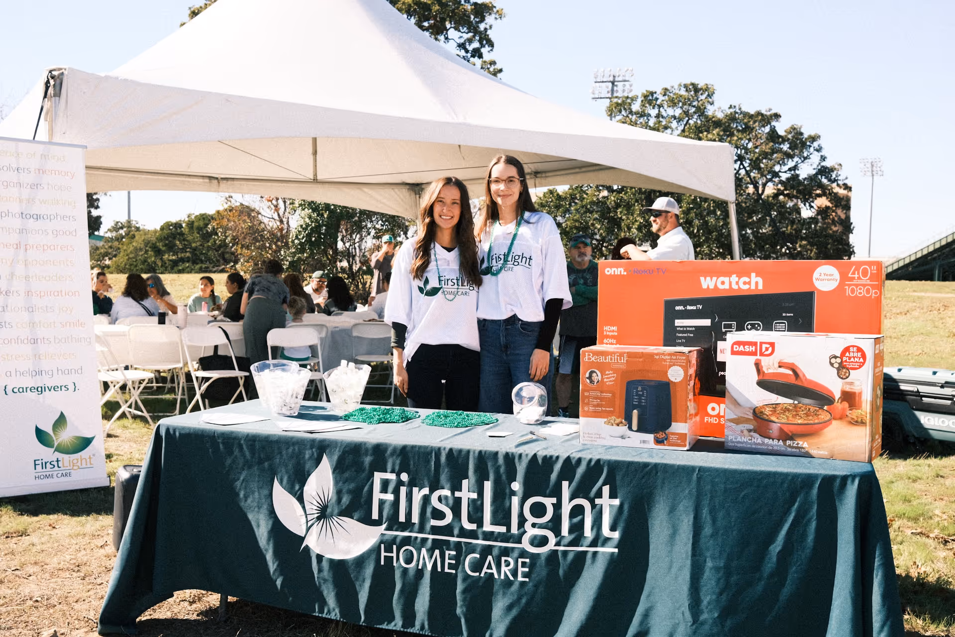 Two staff members stand beside a FirstLight Home Care booth under a white canopy outdoors.