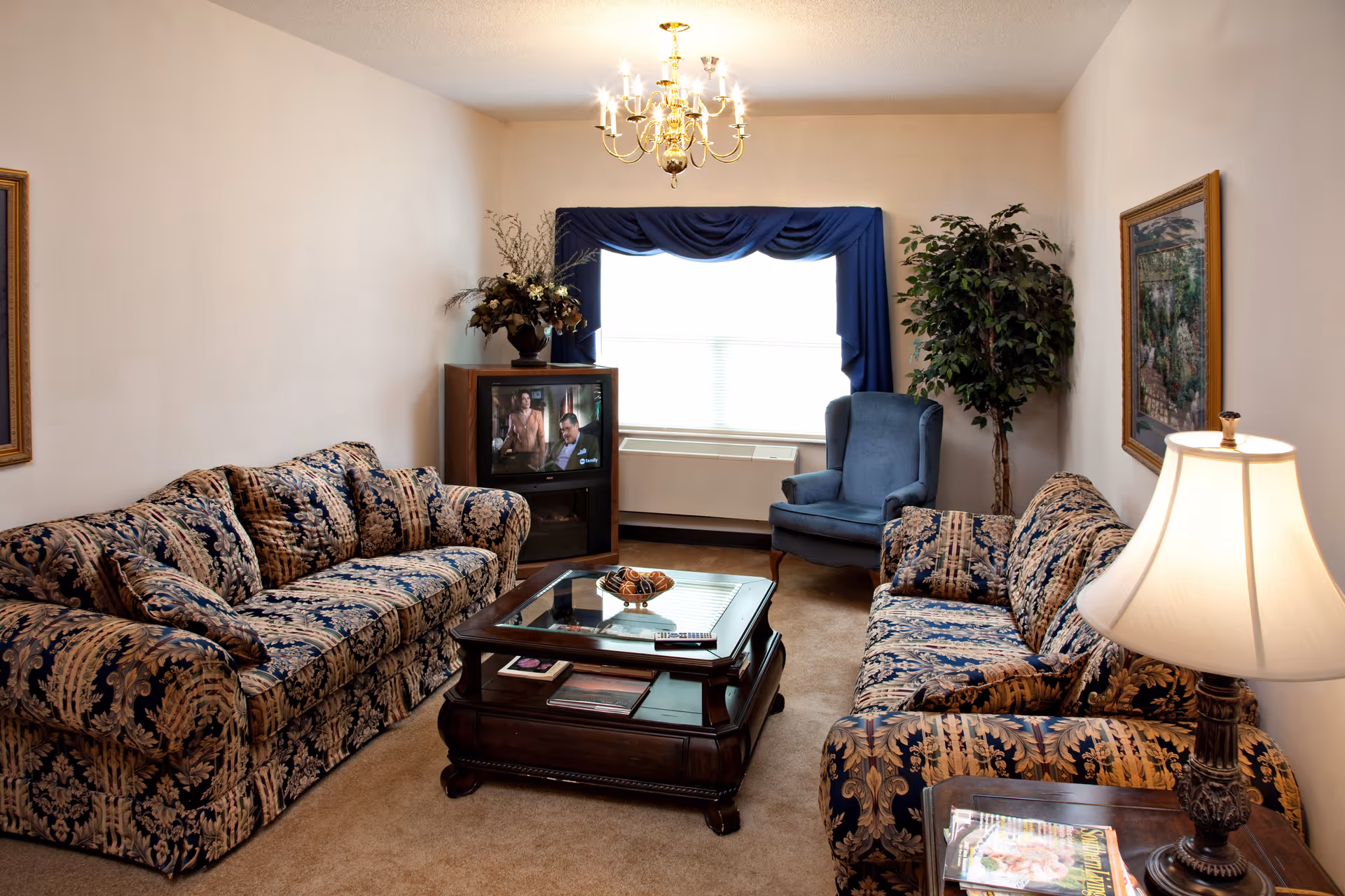 A cozy living room with two patterned sofas facing each other, a wooden coffee table with a glass top in the center, a blue armchair near a window with blue curtains, a TV on a wooden stand, a potted plant, a table lamp on a side table with magazines, and framed artwork on the walls.
