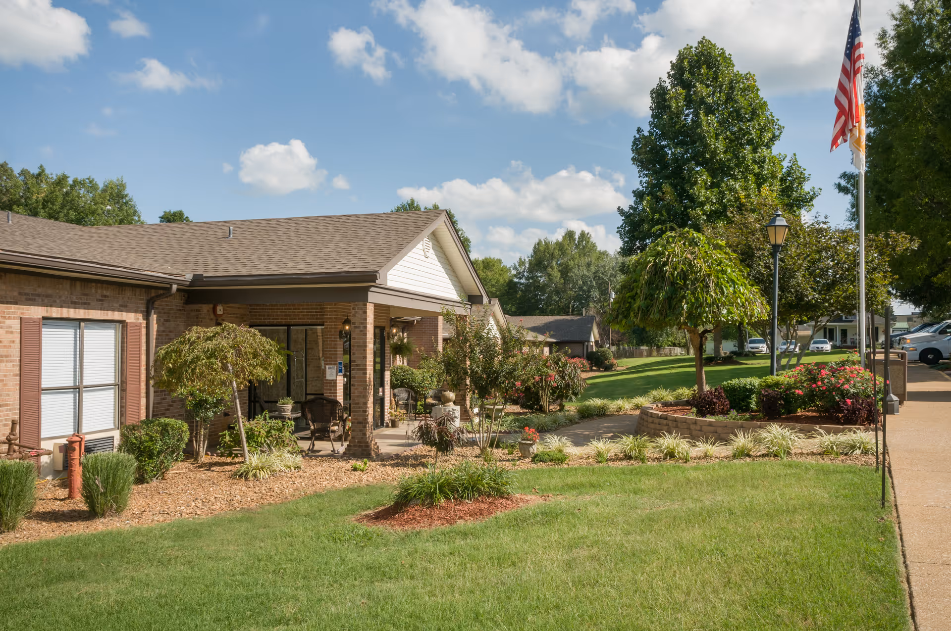 Front exterior of a single-story brick senior living facility with a covered entrance, landscaped lawn, and a flagpole.