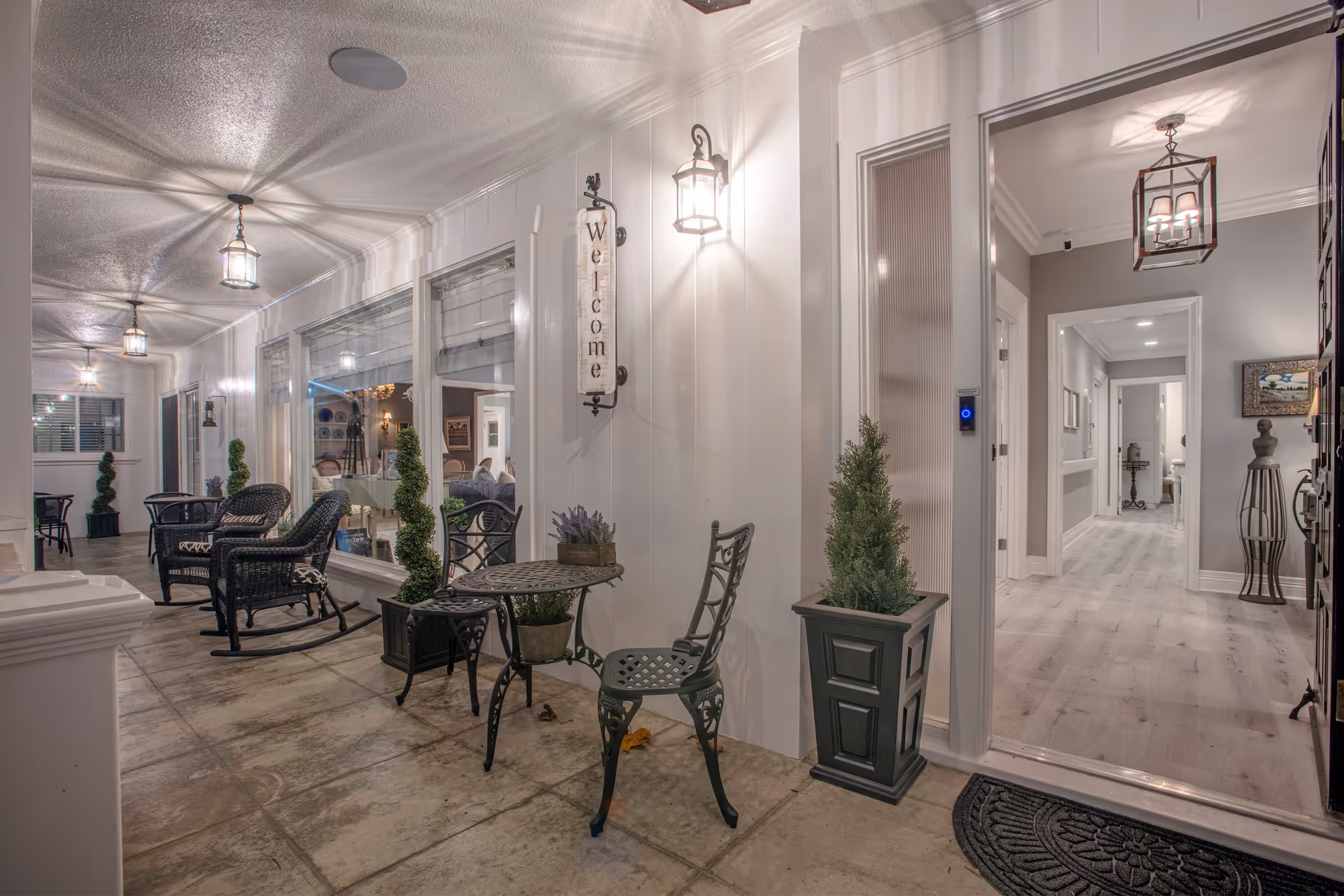 Covered furnished entrance/porch area with chairs, small tables, potted plants, and a 'Welcome' sign leading into a bright hallway.