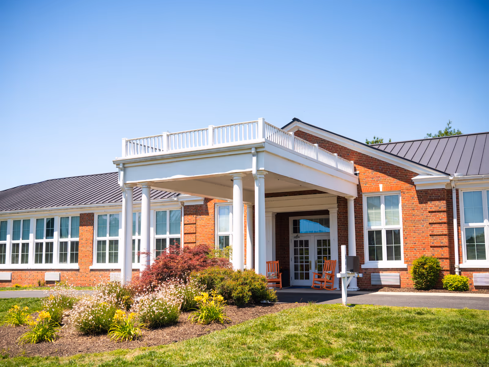 Brick senior living facility with white-columned entry, porch seating, and manicured front yard.