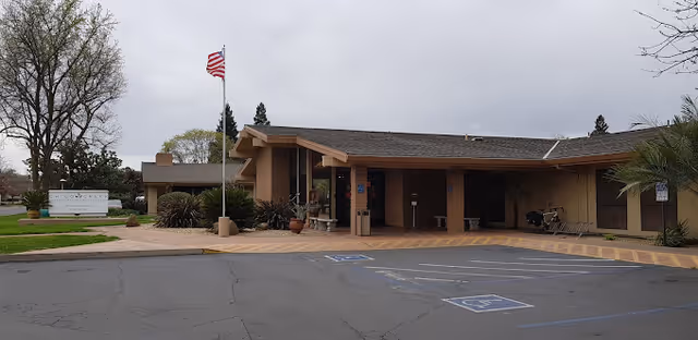 Front entrance of a single-story senior care facility with a covered drive-up, American flag, planters, and marked parking spaces.