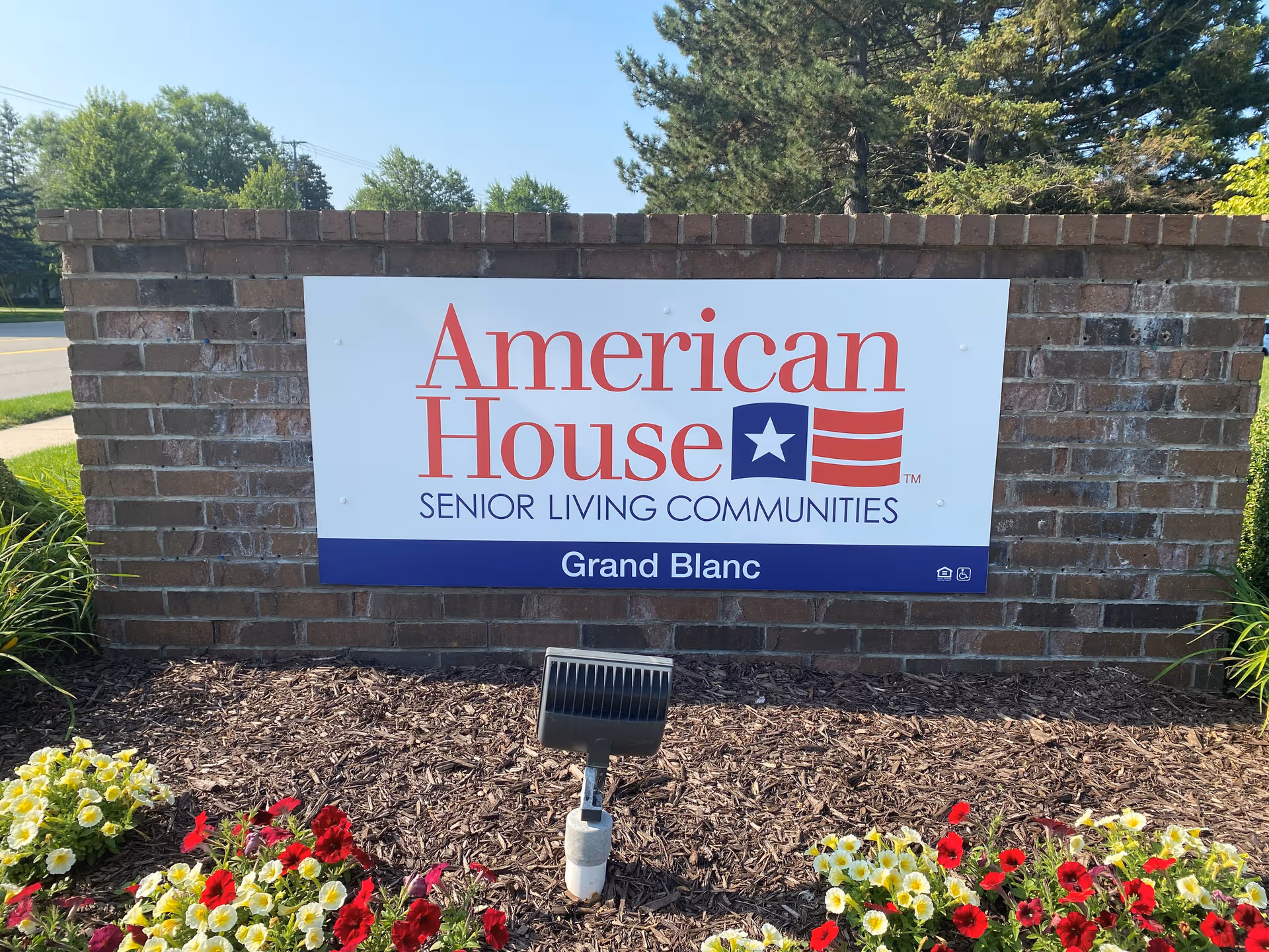 Brick exterior wall sign for American House Grand Blanc with flower beds in front.