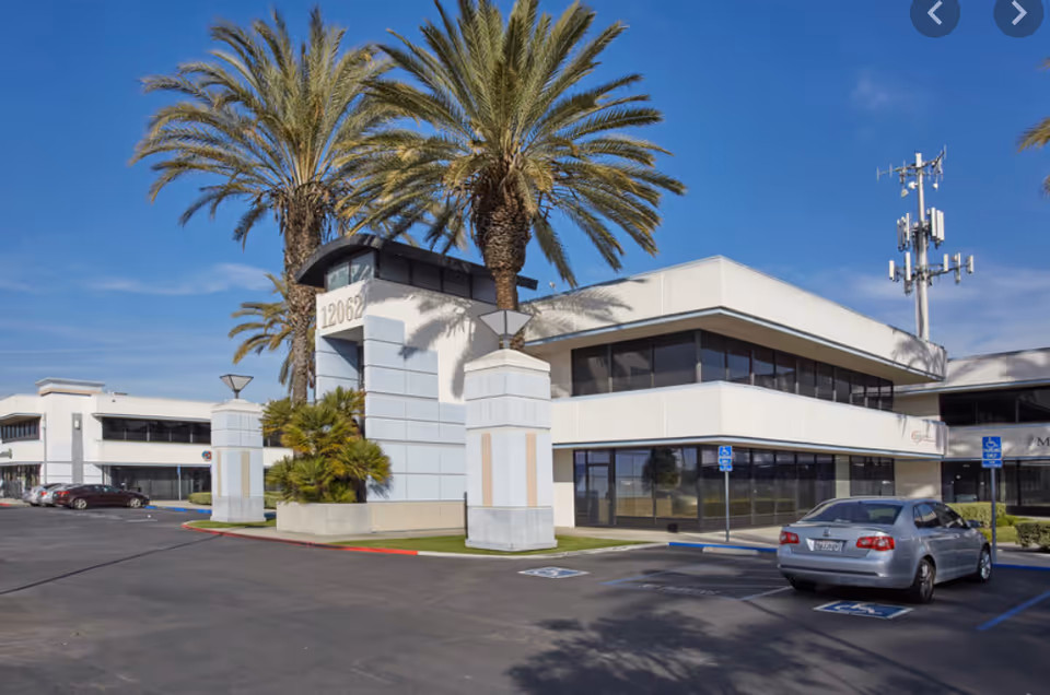 Modern single-story building with glass windows, palm trees, and a parking lot.