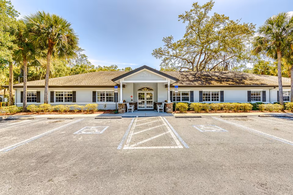 Front exterior view of a single-story building with a covered entrance, surrounded by palm trees and greenery, with a parking lot featuring handicap parking spaces in front.