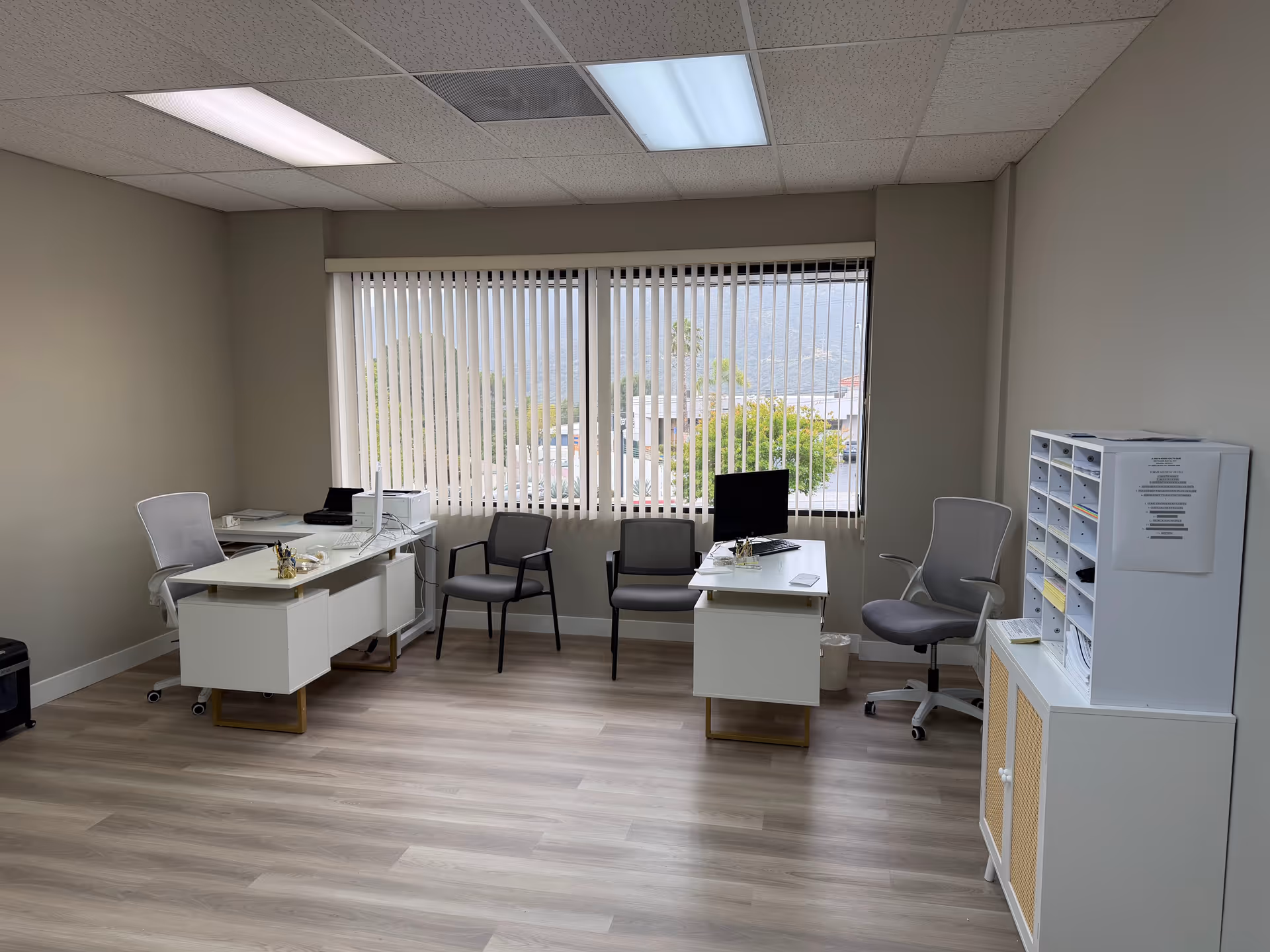 Well-lit office space with desks, chairs, and filing cabinets by a window.