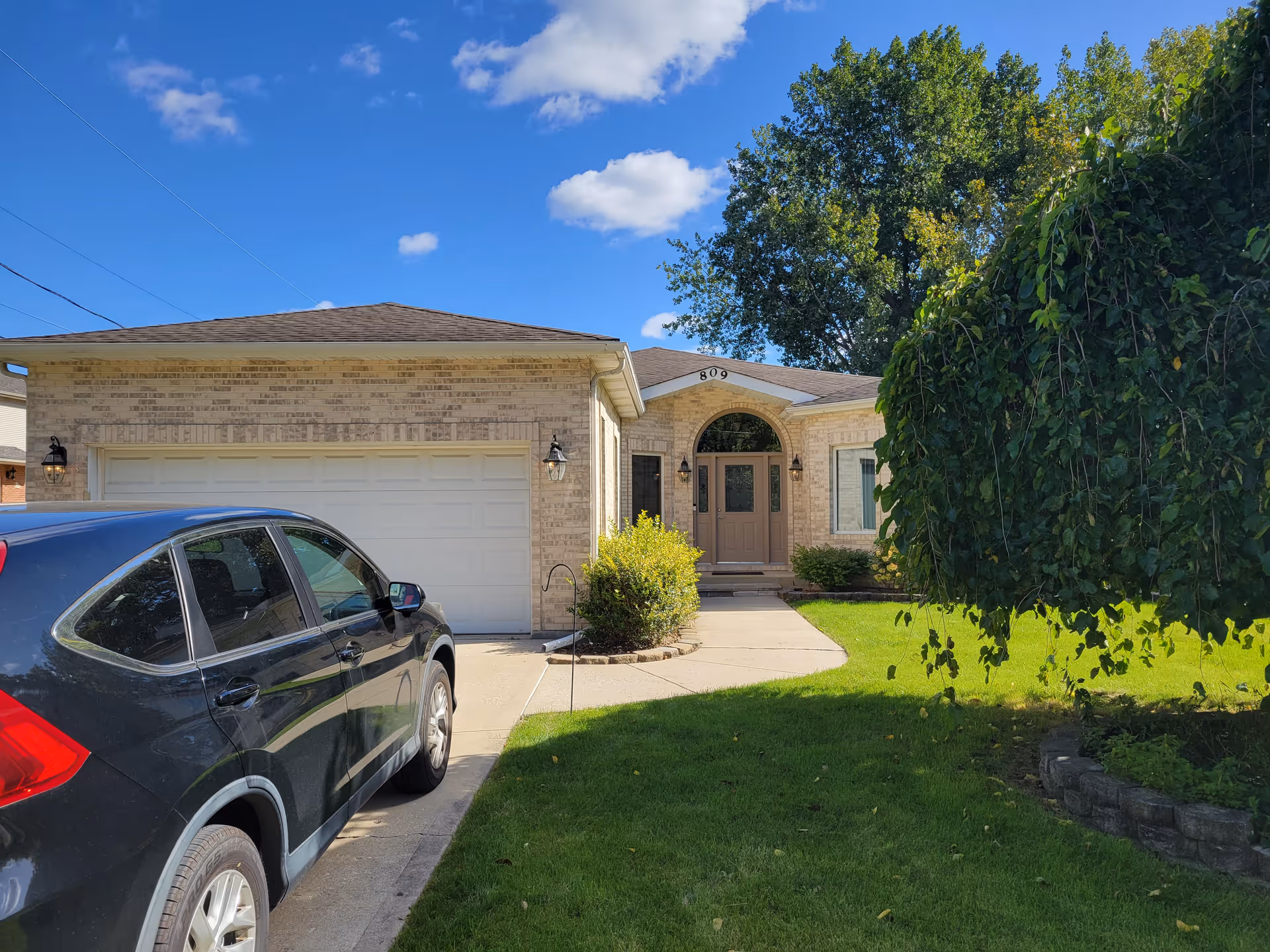 Front view of a single-story brick home with a two-car garage, a car in the driveway, and a green lawn with trees.