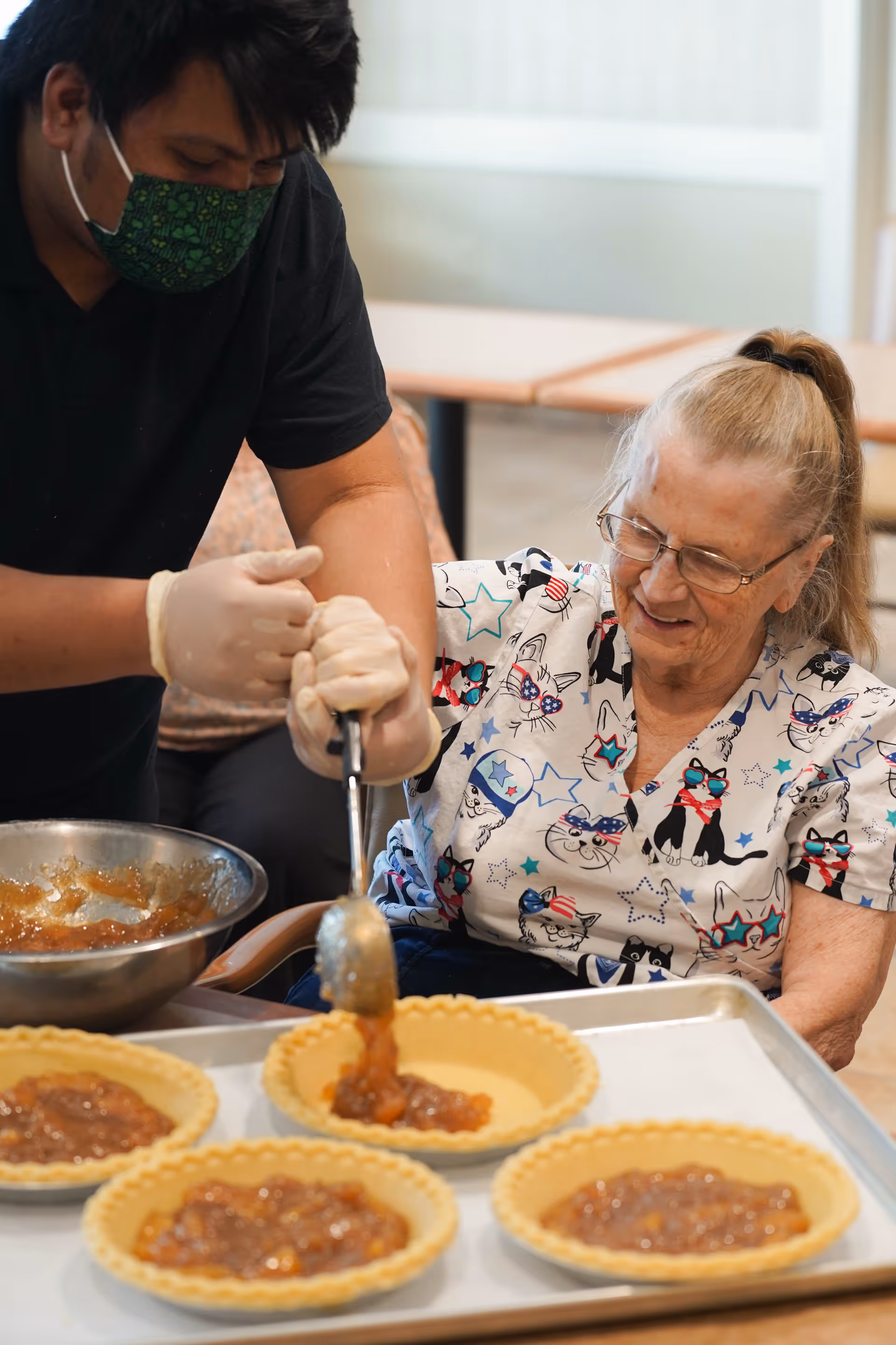 A caregiver wearing a green patterned face mask and gloves helps an elderly woman with glasses and a cat-themed shirt spoon filling into pie crusts on a baking tray in a kitchen or activity area.
