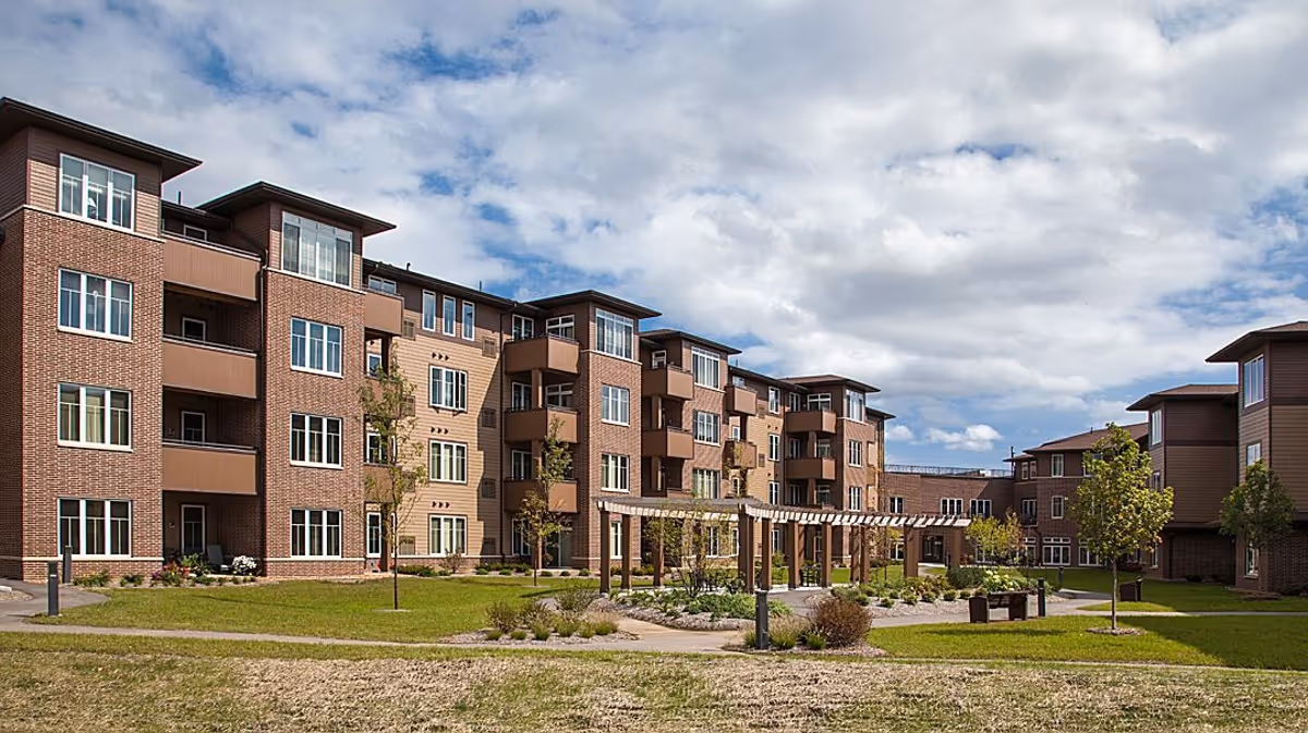 Exterior view of a multi-story senior living facility with brick and beige siding, featuring balconies, windows, a landscaped courtyard with a pergola, trees, and walking paths under a partly cloudy sky.
