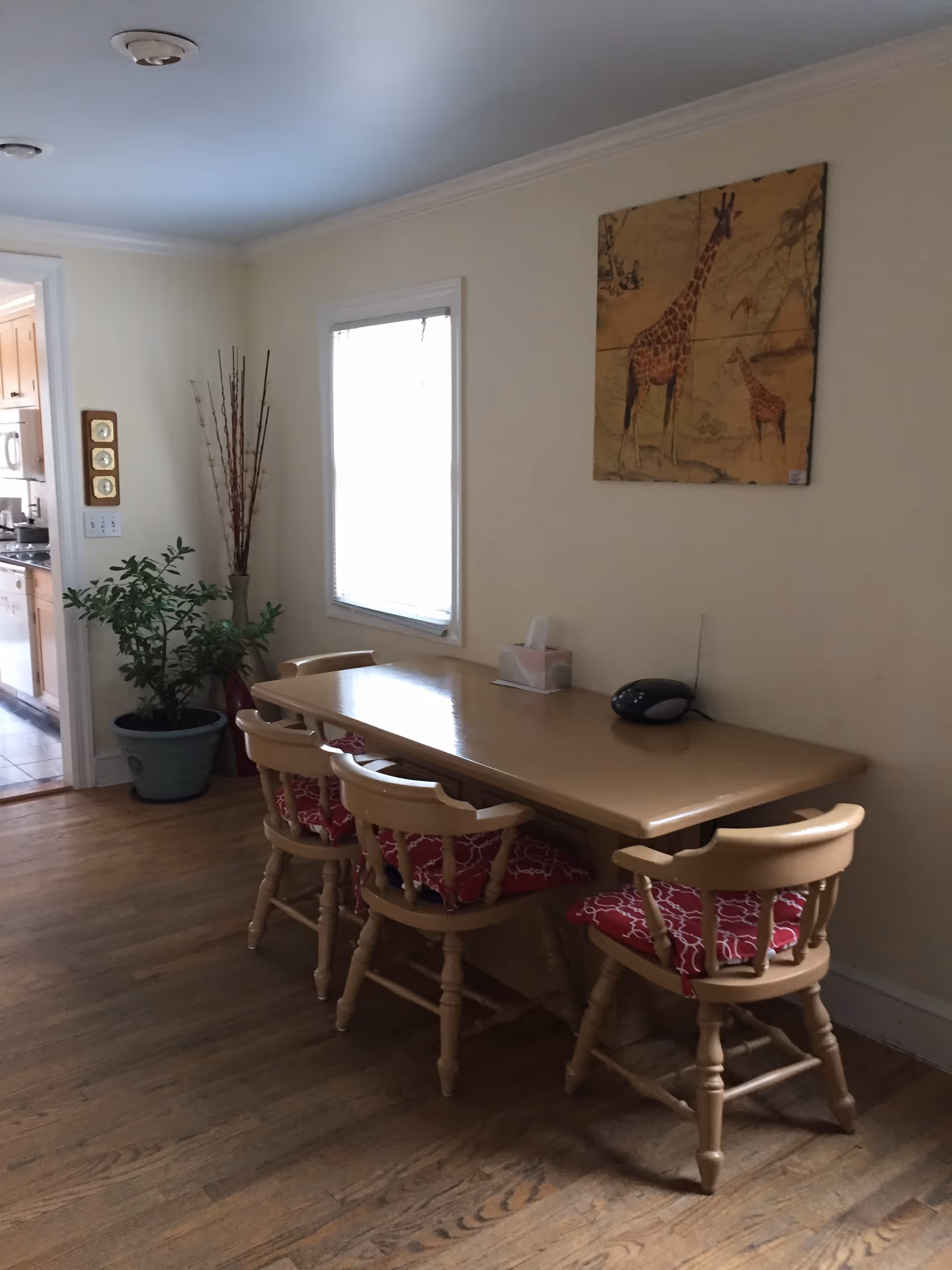 Wood dining table with four chairs, red cushions, plants, and giraffe wall art.