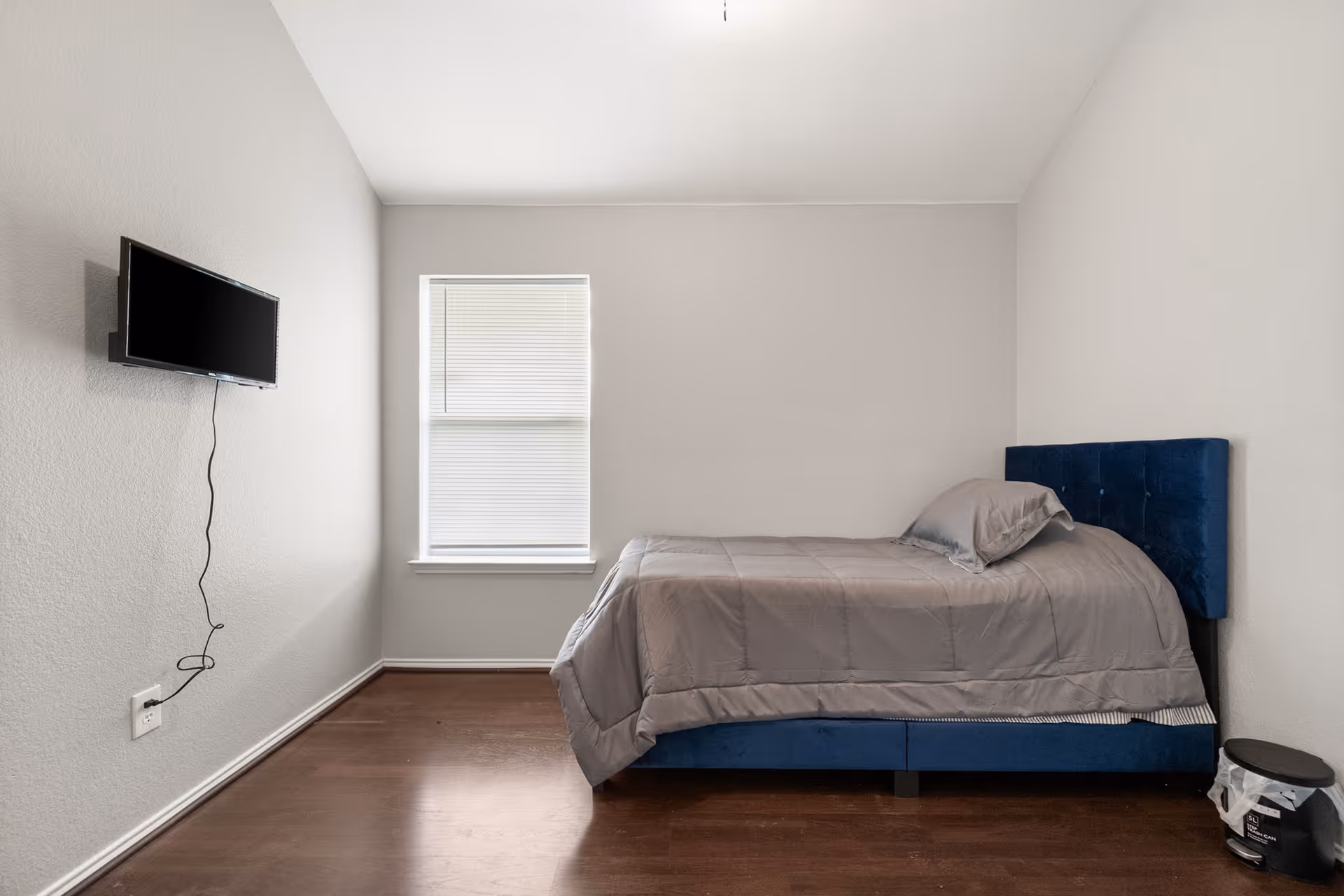 A small minimalist bedroom with a blue upholstered bed, gray bedding, window, and wall-mounted TV.