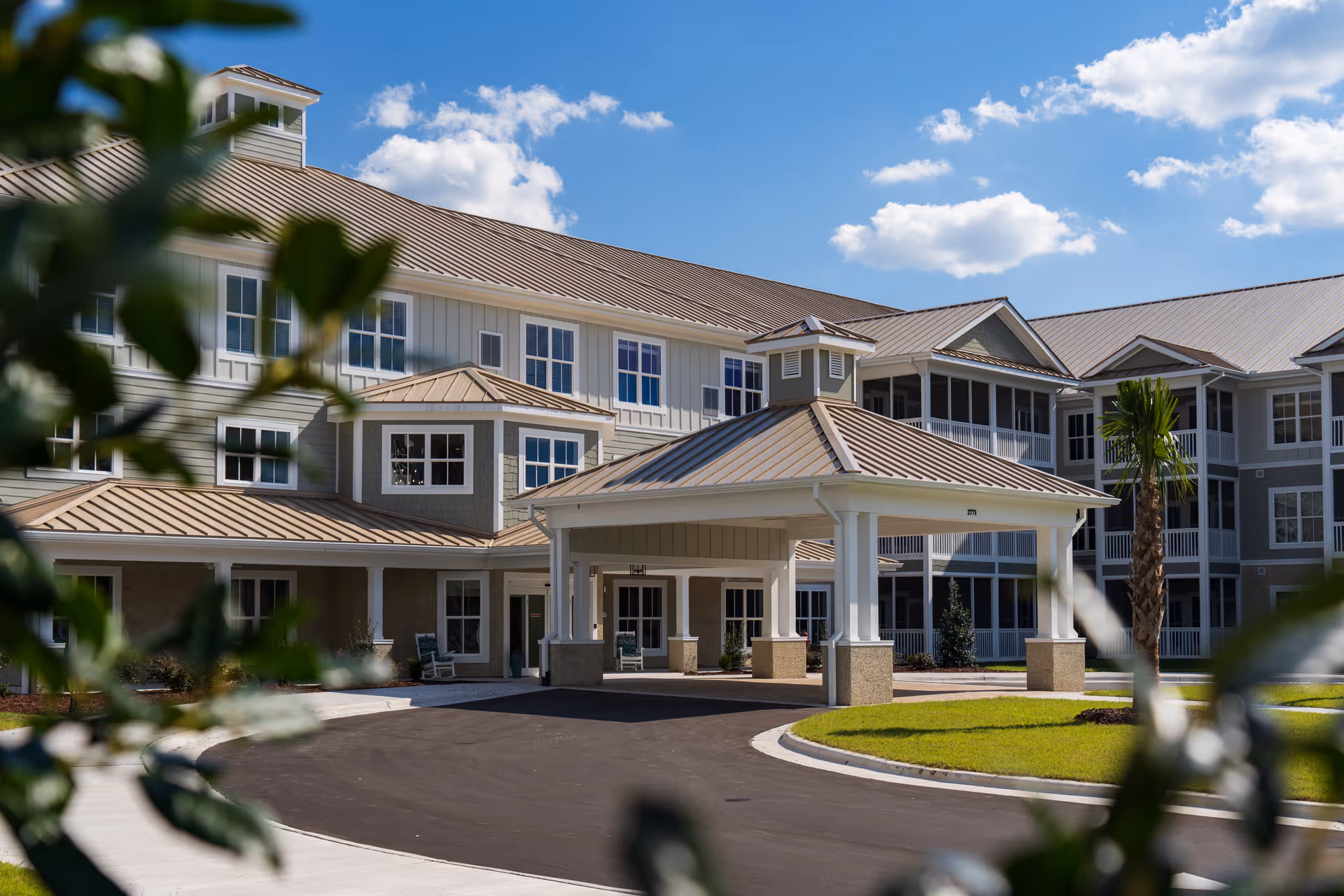 Exterior view of a senior living facility building with a covered entrance, multiple windows, and a metal roof under a blue sky with scattered clouds. There are some green plants and a palm tree in the foreground.