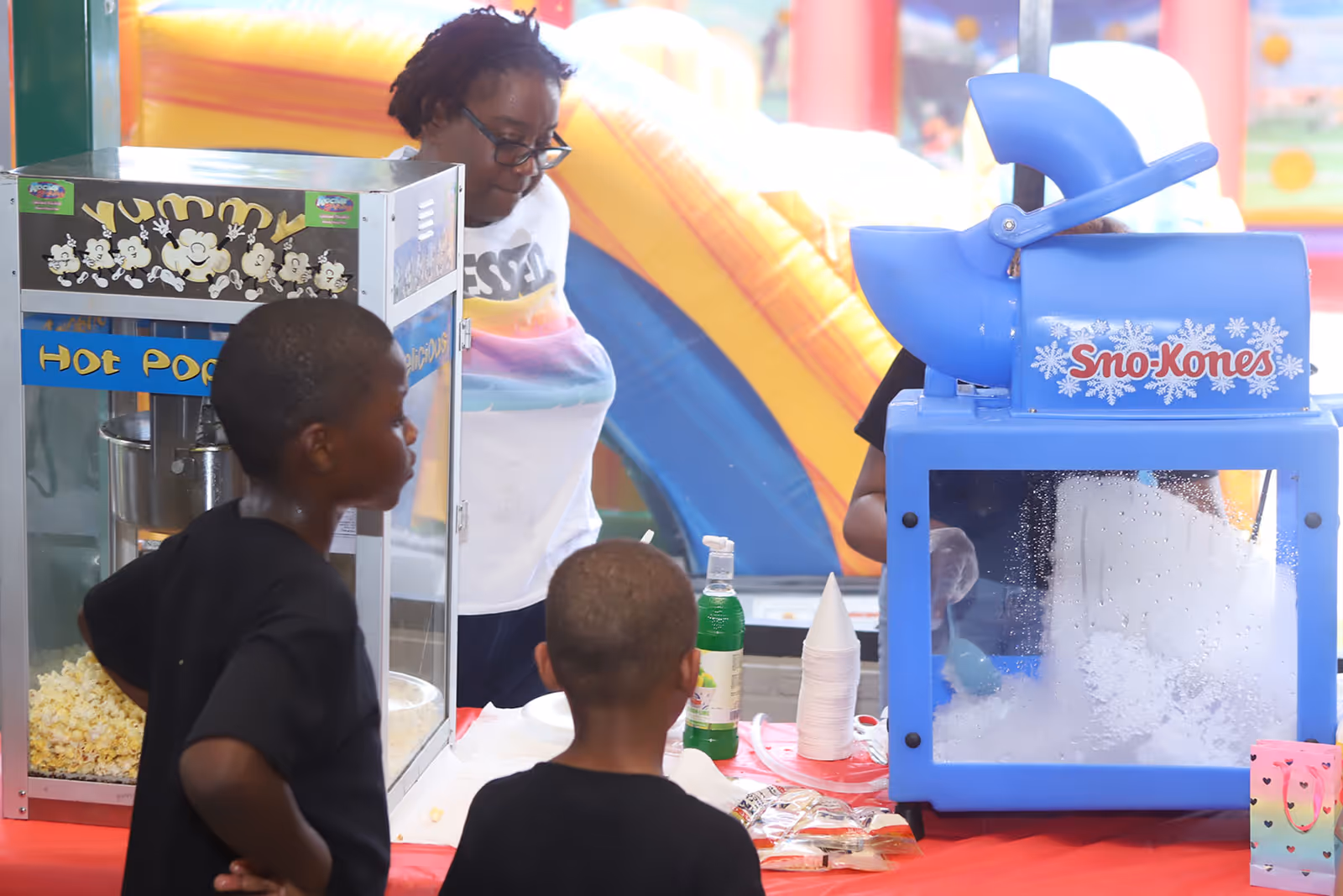 A staff member serves popcorn while kids enjoy a Sno-Kone machine at a colorful indoor event.