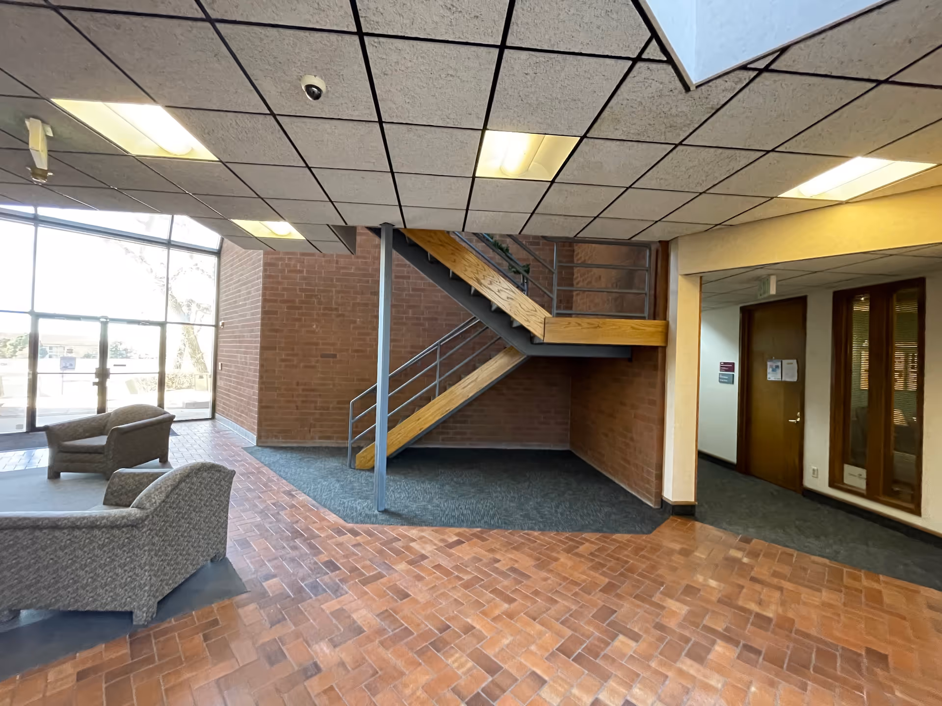 Lobby area with brick walls, a wooden staircase, and seating near large glass doors.