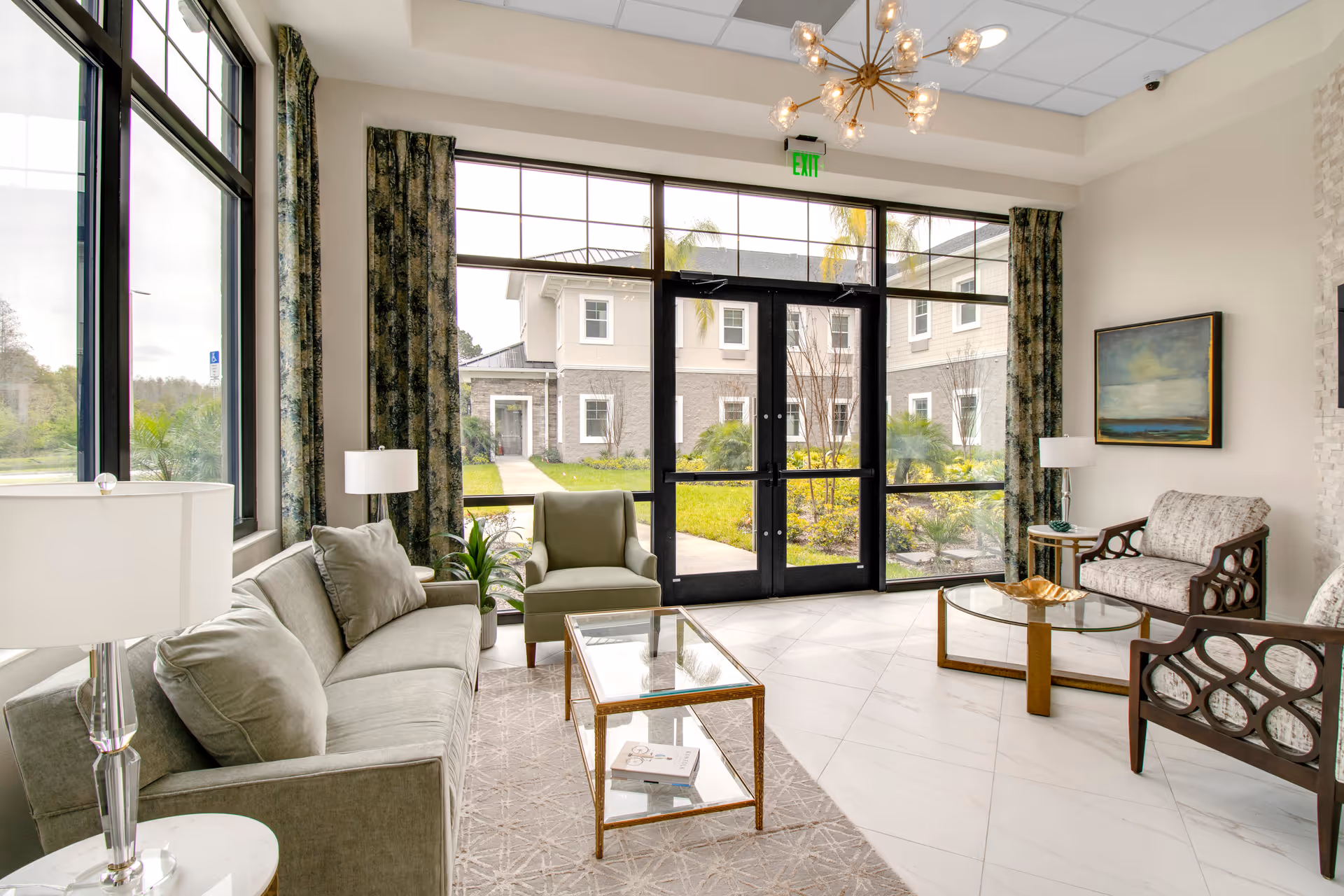Bright and modern living room area with large windows and glass doors leading outside. The room features a gray sofa with matching pillows, an armchair, two patterned armchairs, a glass coffee table, side tables with lamps, and a decorative chandelier. There is a painting on the wall and green curtains framing the windows.