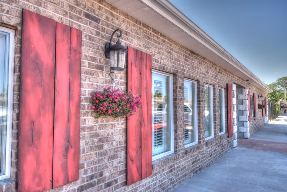 Exterior view of a brick building with several windows, each framed by red wooden shutters. There are hanging flower baskets with pink flowers and black outdoor lanterns mounted on the wall. The sidewalk runs alongside the building under a clear blue sky.