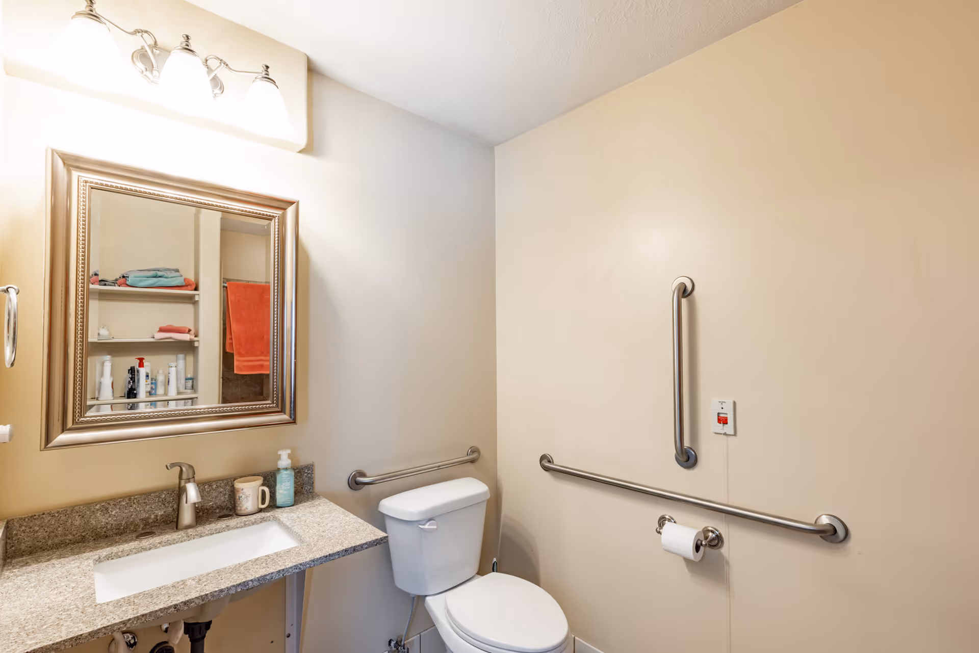 Accessible bathroom with sink, mirror, toilet, grab bars, and toiletries on a granite counter.