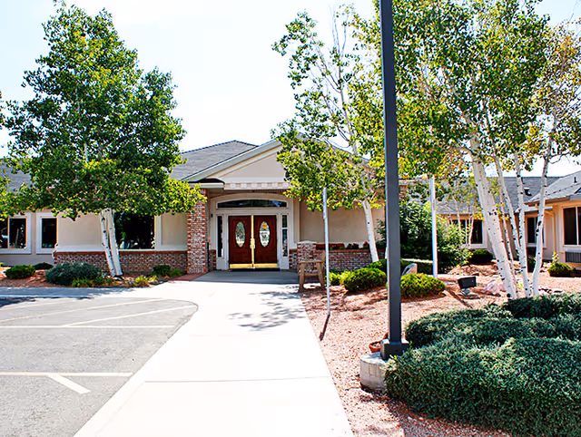 Exterior view of Willow Wind Assisted Living facility showing the entrance with double wooden doors, surrounded by trees and landscaped bushes, with a sidewalk and parking area in front.