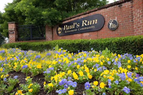 A brick wall with a sign that reads 'Paul's Run Retirement Community' surrounded by green bushes and a flower bed with yellow and purple flowers in front. There is also a gate and trees in the background.