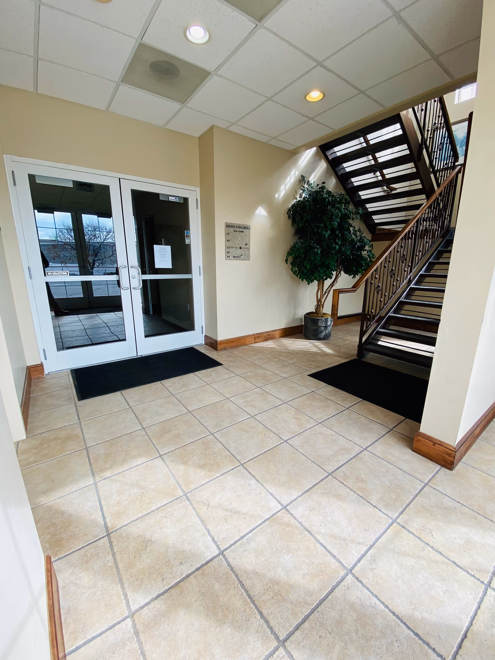 Lobby with double glass doors, tiled floor, staircase, and a potted plant.