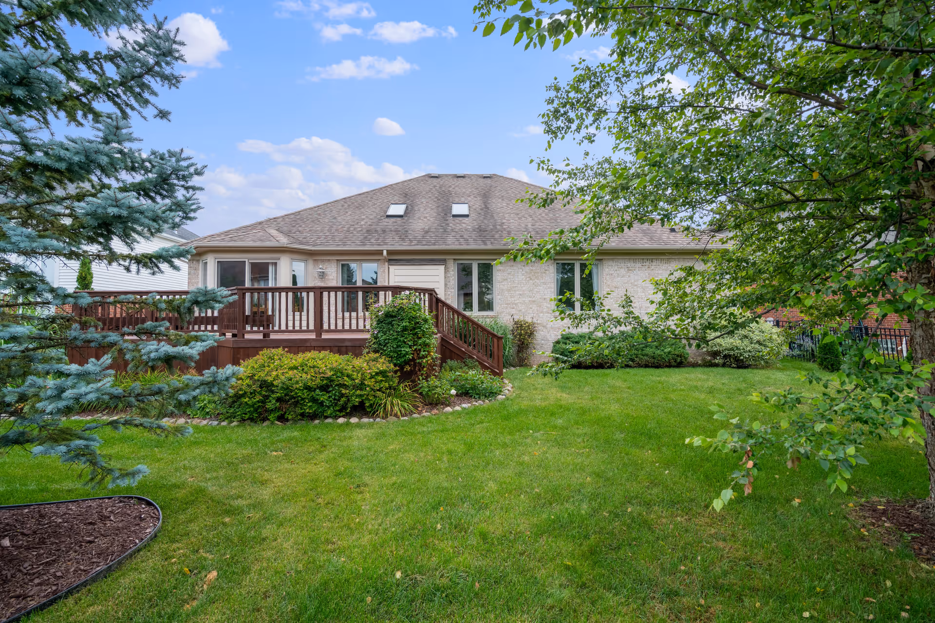 Backyard view of a single-story house with a wooden deck, surrounded by green grass, bushes, and trees under a partly cloudy sky.