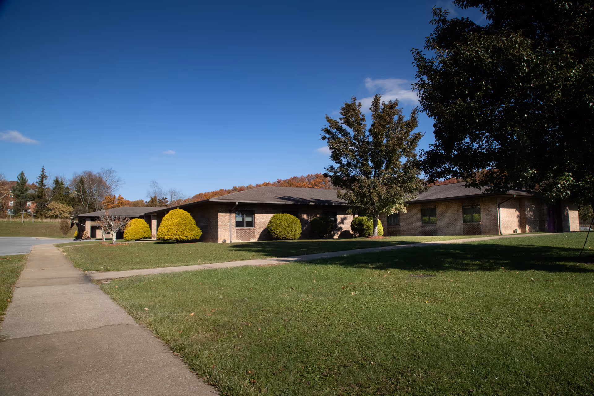 A brick senior living facility with a manicured lawn and trees under a clear blue sky.