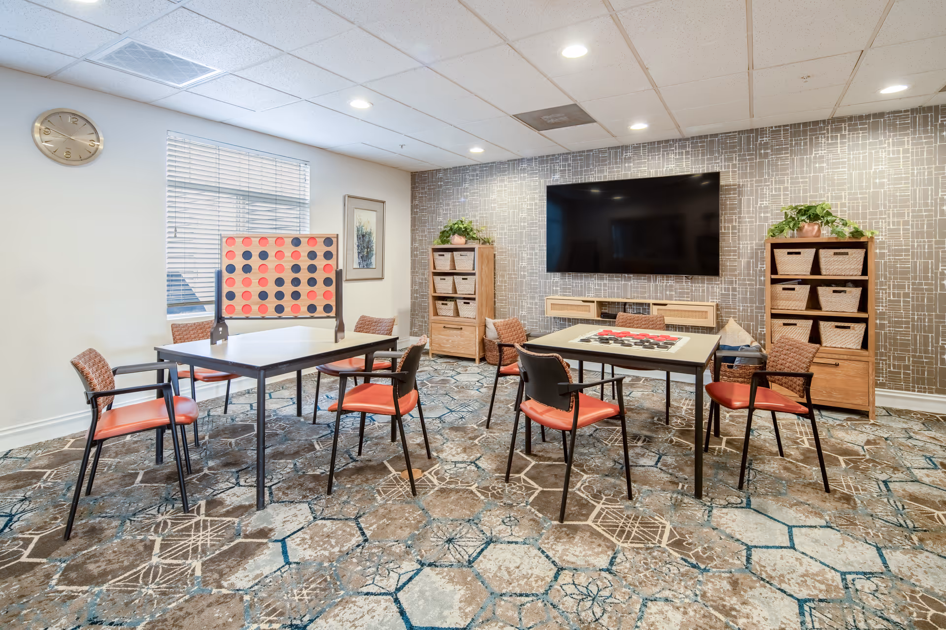 Communal game room with tables, chairs, shelves, and a wall-mounted TV.