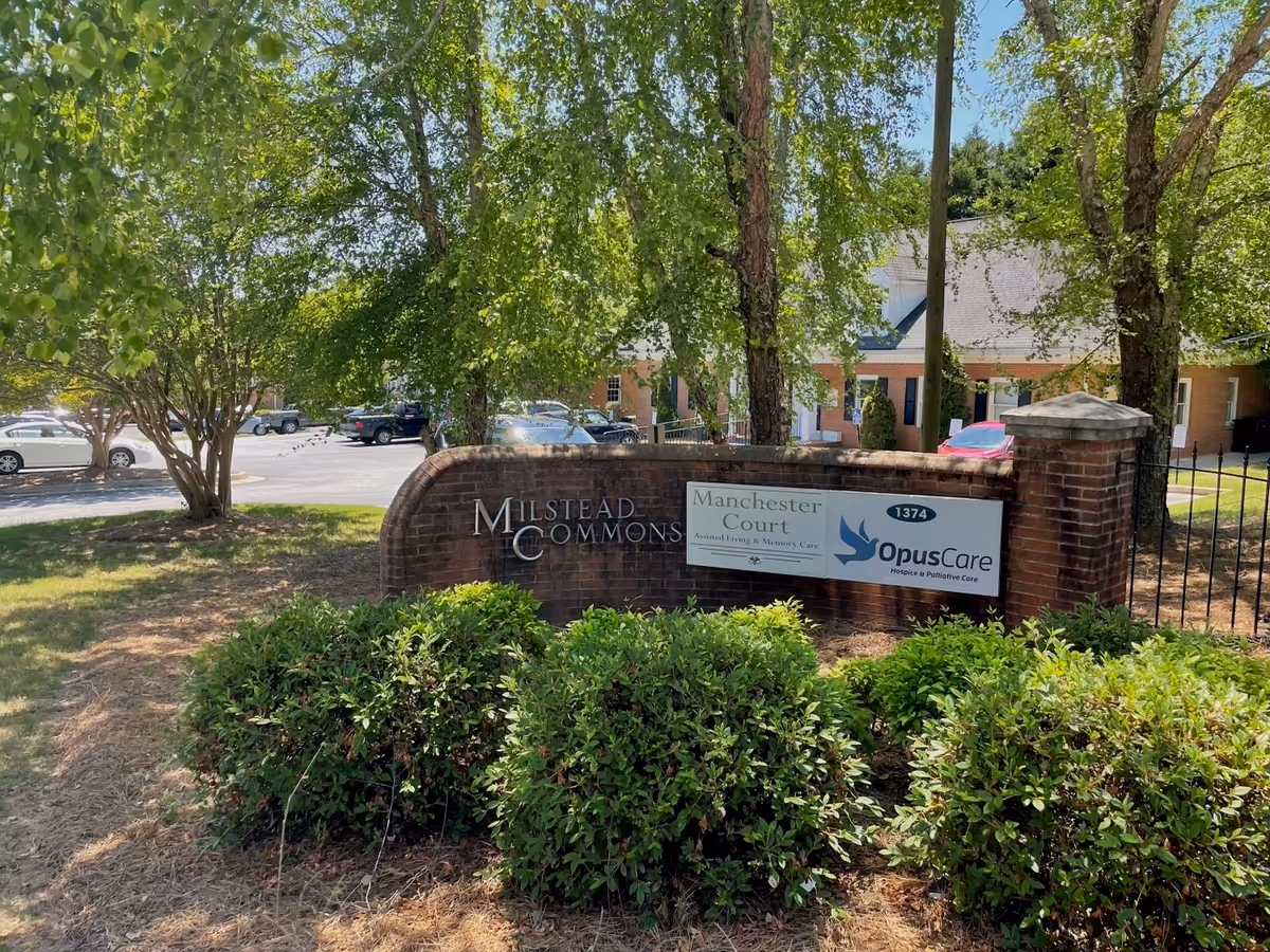 Brick entrance sign for Milstead Commons and OpusCare with trees and parked cars nearby.